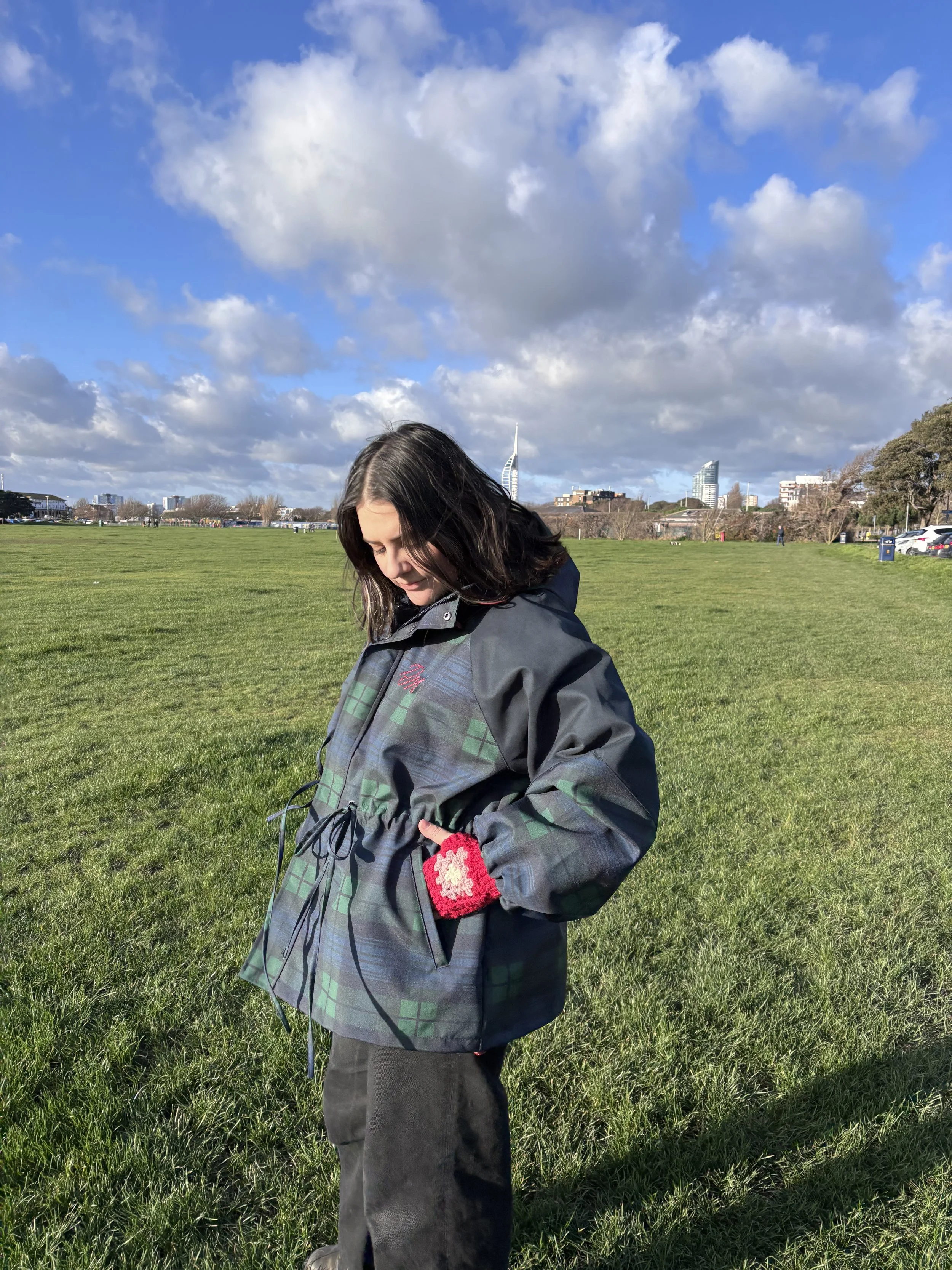 A young woman standing in a grassy park on a sunny day with clouds in the sky, wearing a plaid jacket and red crocheted gloves.