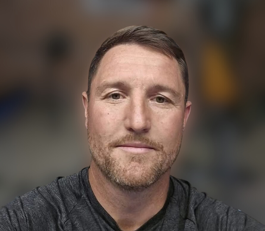 A close-up portrait of a man with short brown hair, a beard, and wearing a dark gray shirt, against a blurred background.
