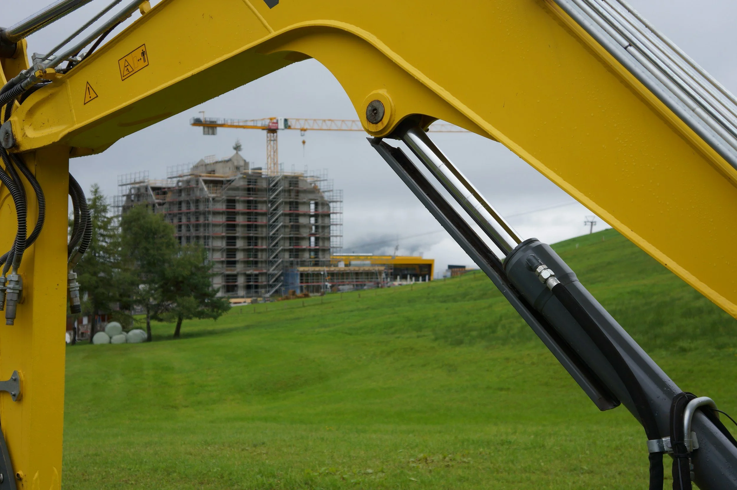 Close-up of a yellow construction excavator arm with hydraulic piston, with a building under construction and a crane in the background on a cloudy day.