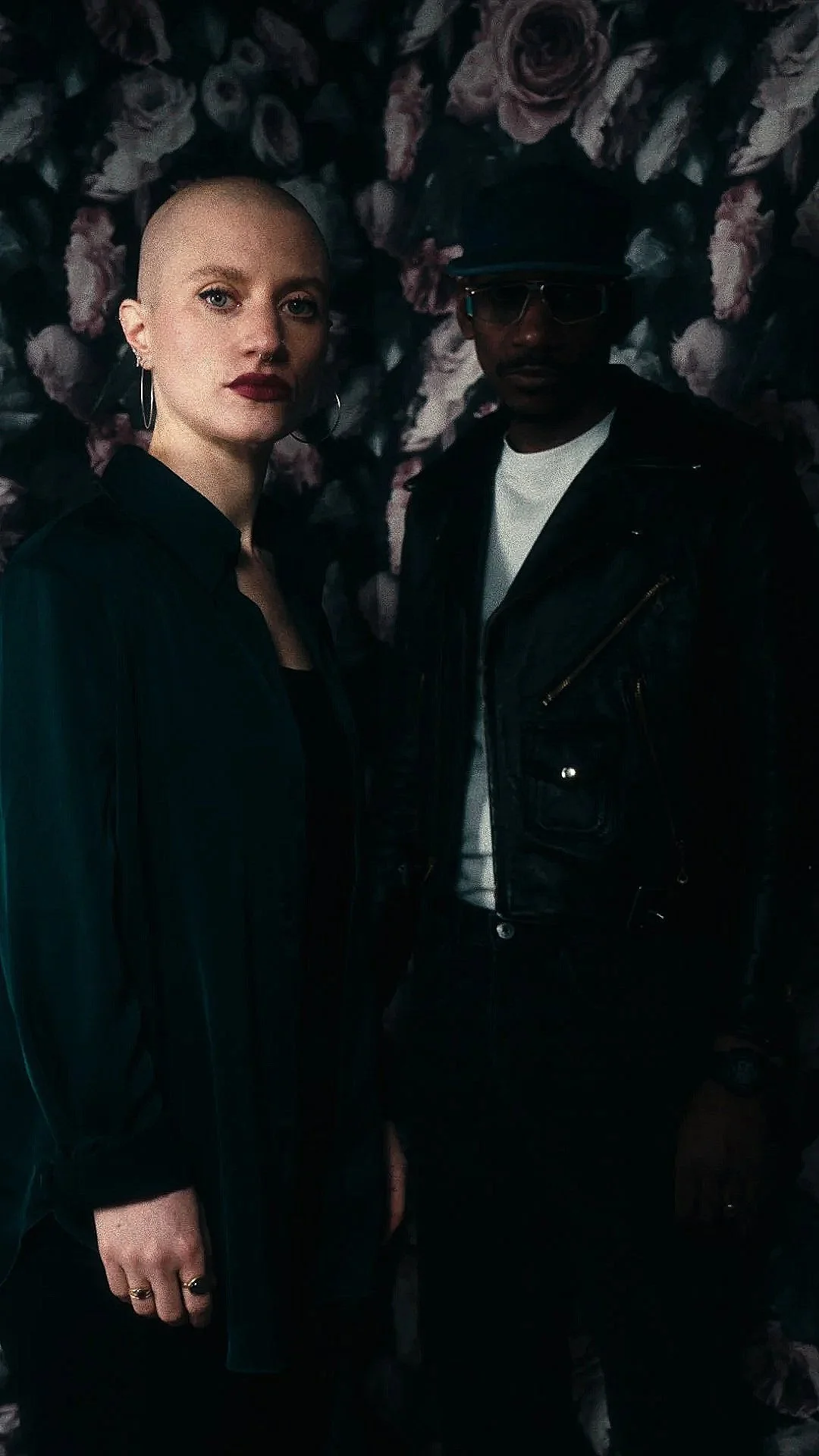 A white woman (Leanor) with a shaved head and wearing a dark green shirt and gold hoop earrings is standing next to a black man wearing vintage sunglasses, a dark blue hat, and a fitted leather biker jacket, against a background of large flowers.