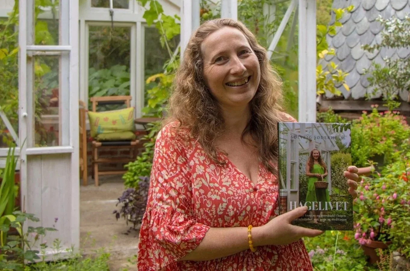 Kvinne som smiler og holder en bok i en hage med grønne planter og blomster.