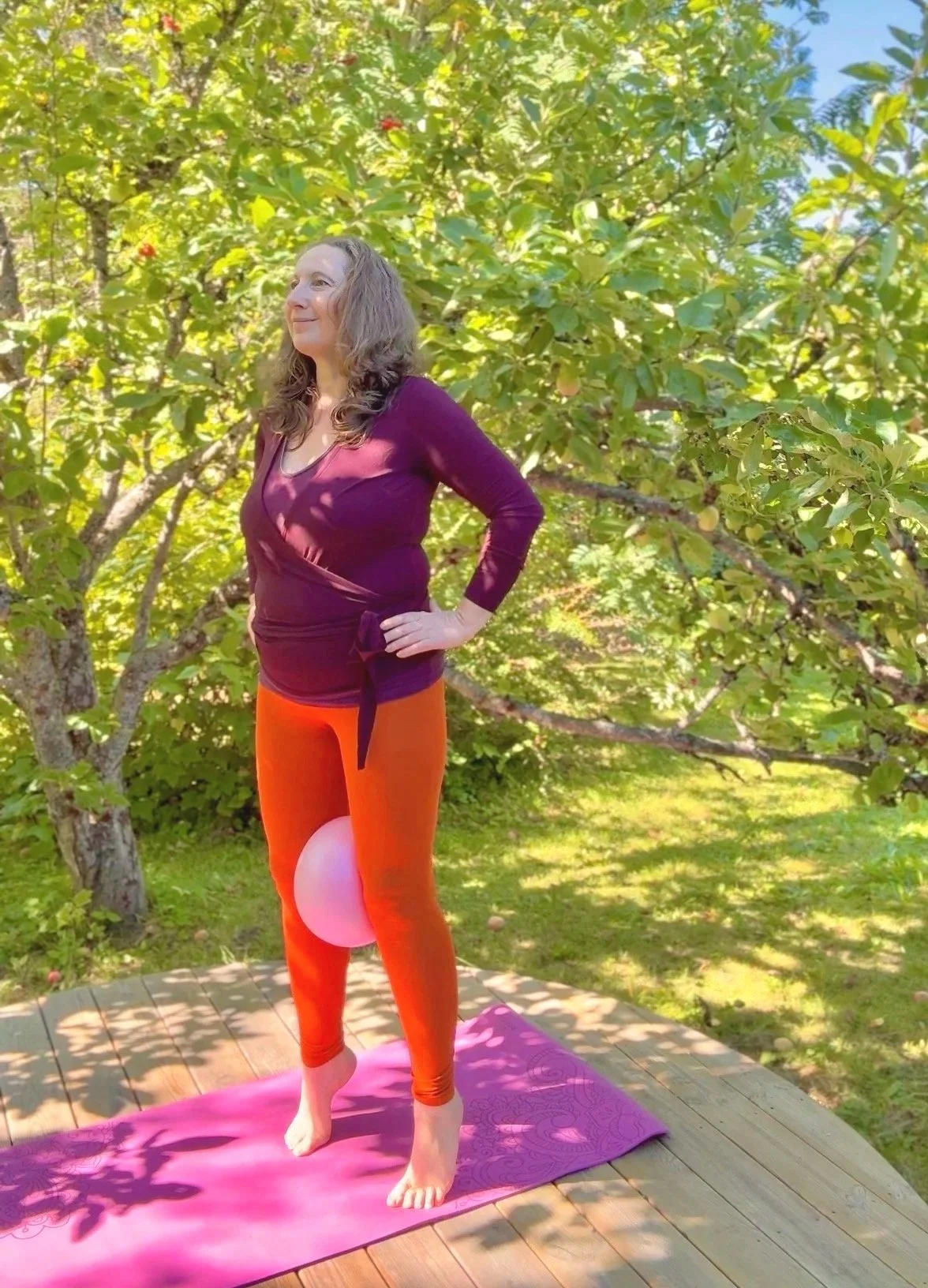 en kvinne gjør bekkenbunnstrening med ball ute i hagen på en yogamatte