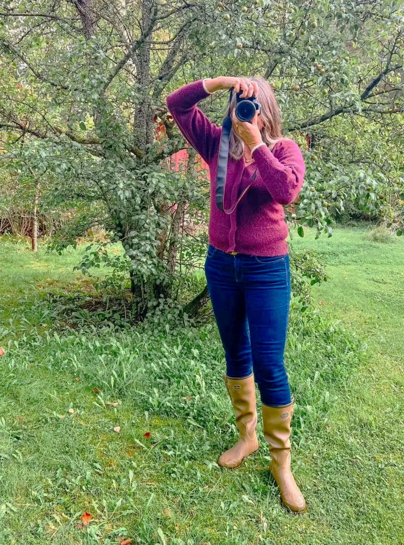 Person som tar et bilde i en hage med trær og grønt gress, iført beige støvler, blå jeans og en rød/genser.