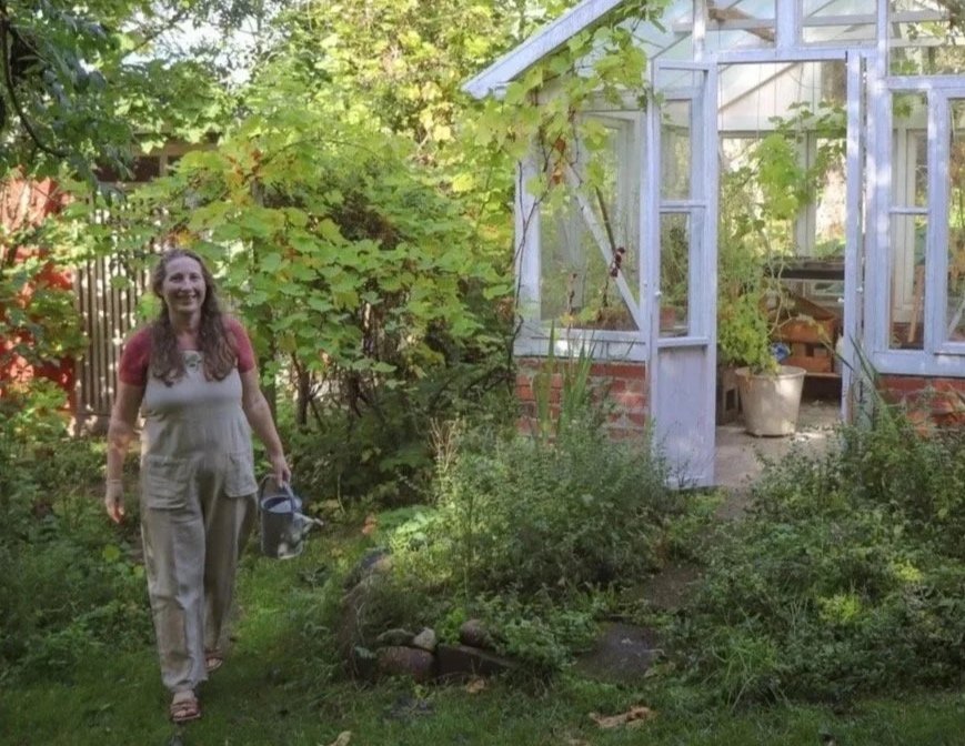 En kvinne i hage med grønnsaker og et lite drivhus i bakgrunnen.