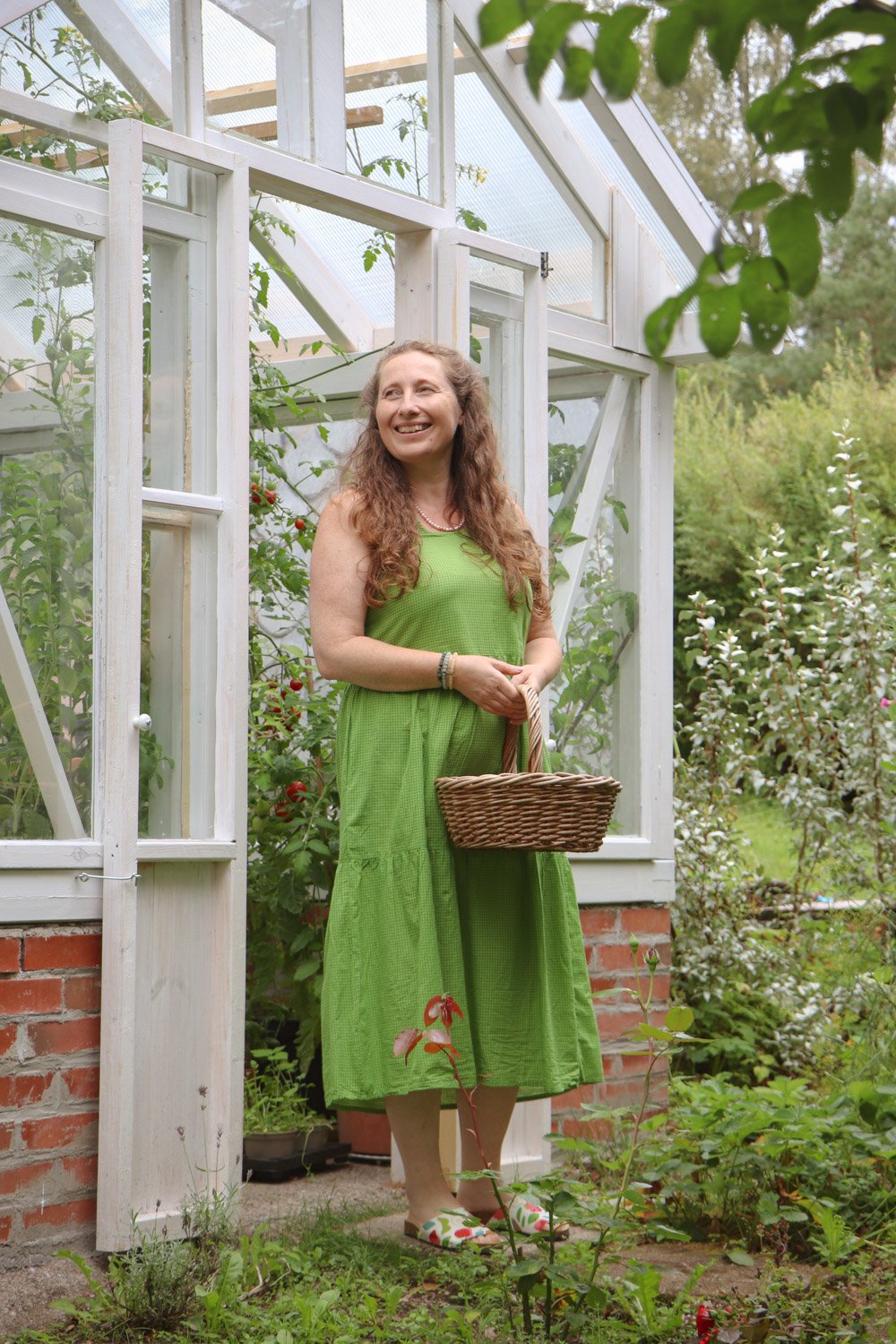 Kvinne med kurv plukker ferske grønnsaker foran drivhuset i hagen.