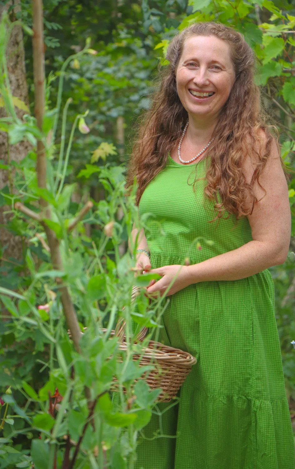 Kvinne som holder en grønnsakskurv ute i hagen foran drivhuset.