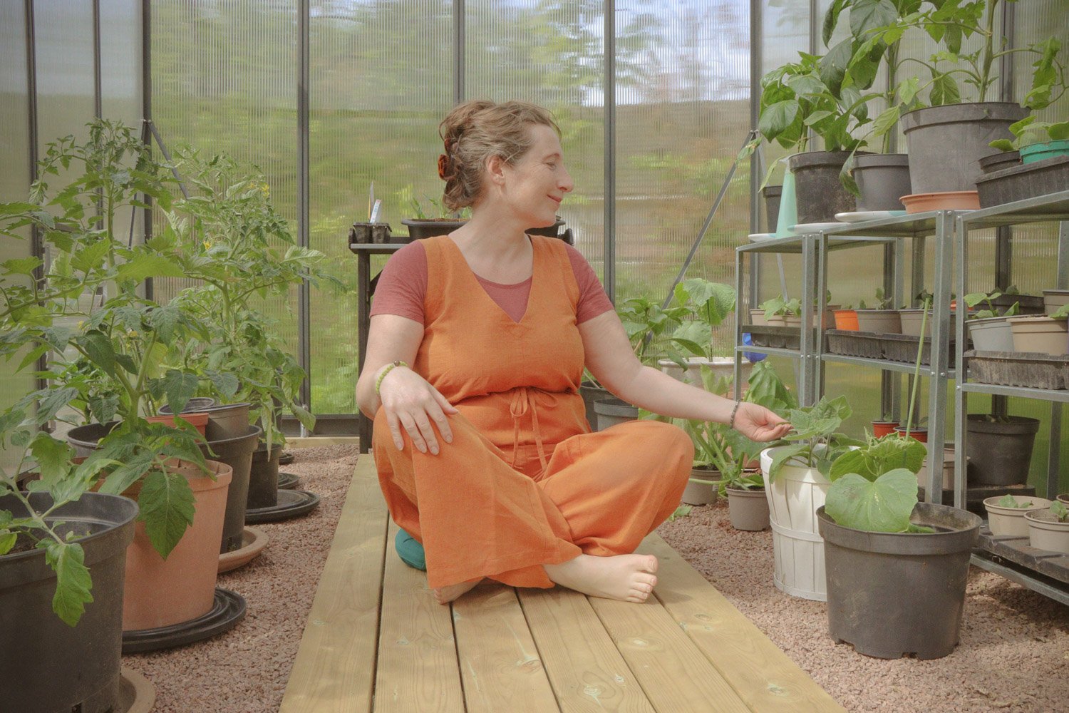 Kvinne sitter i yogaposisjon på gulvet i drivhuset og berører et planteblad, rolig og harmonisk