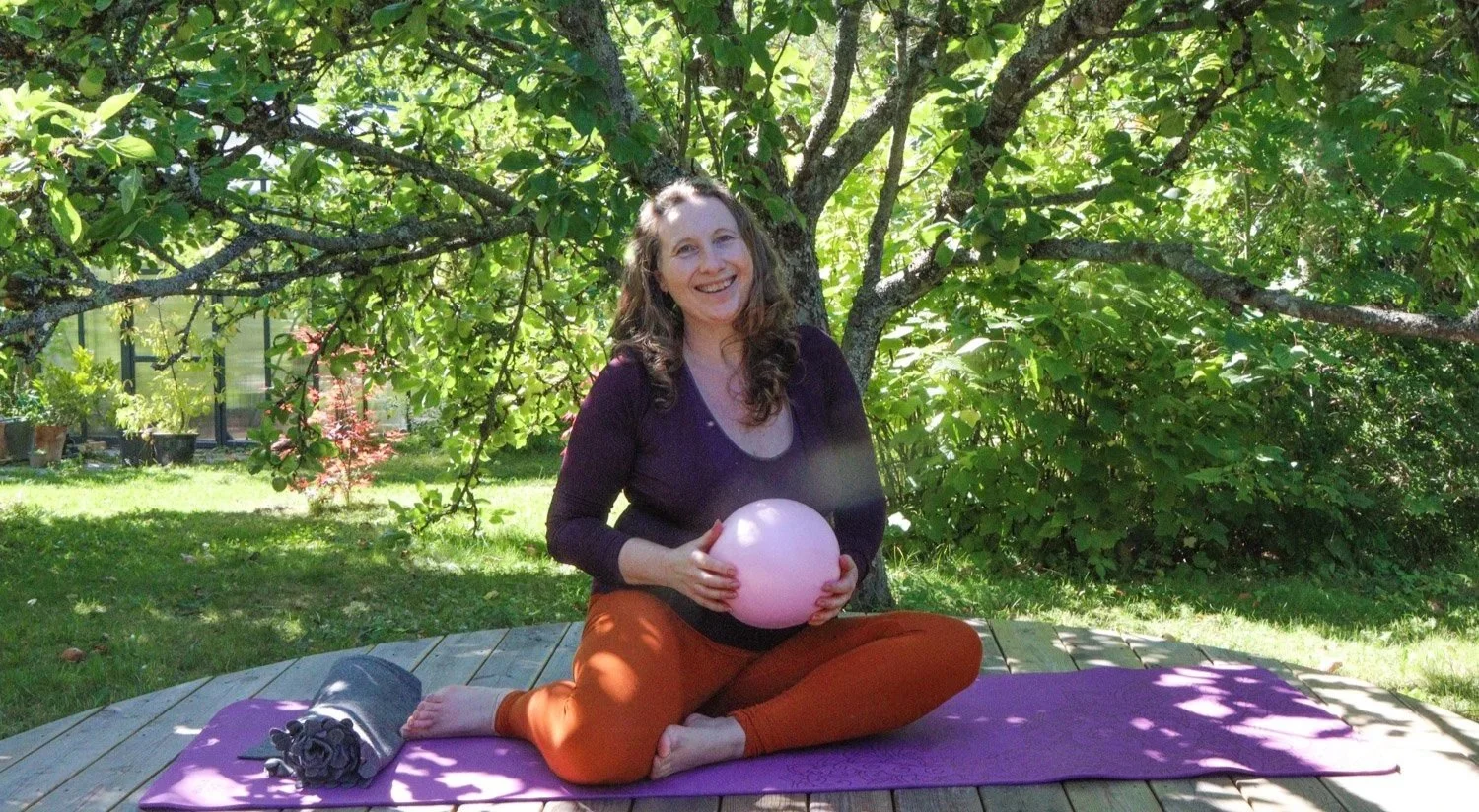 Kvinne som sitter på en yogamatte i en hage med grønne trær og planter, holder en rosa ball, smiler og har på seg oransje bukser og en mørk genser.