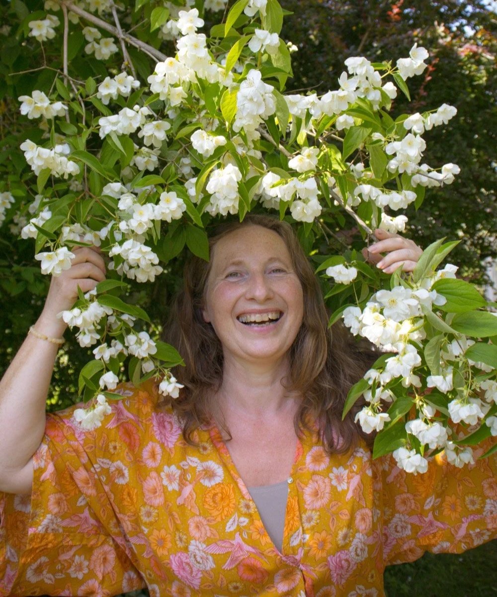 Kvinne i hagen holder duftsjærsminbusker, ler og finner ro med yoga og natur. Opplev stressfrihet, glede og velvære ute.