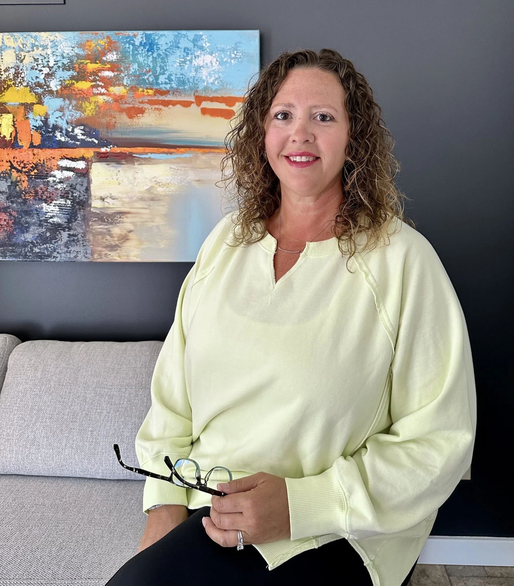 A woman with curly hair wearing a light yellow sweatshirt, sitting on a beige couch, holding glasses, in front of an abstract landscape painting on a gray wall.
