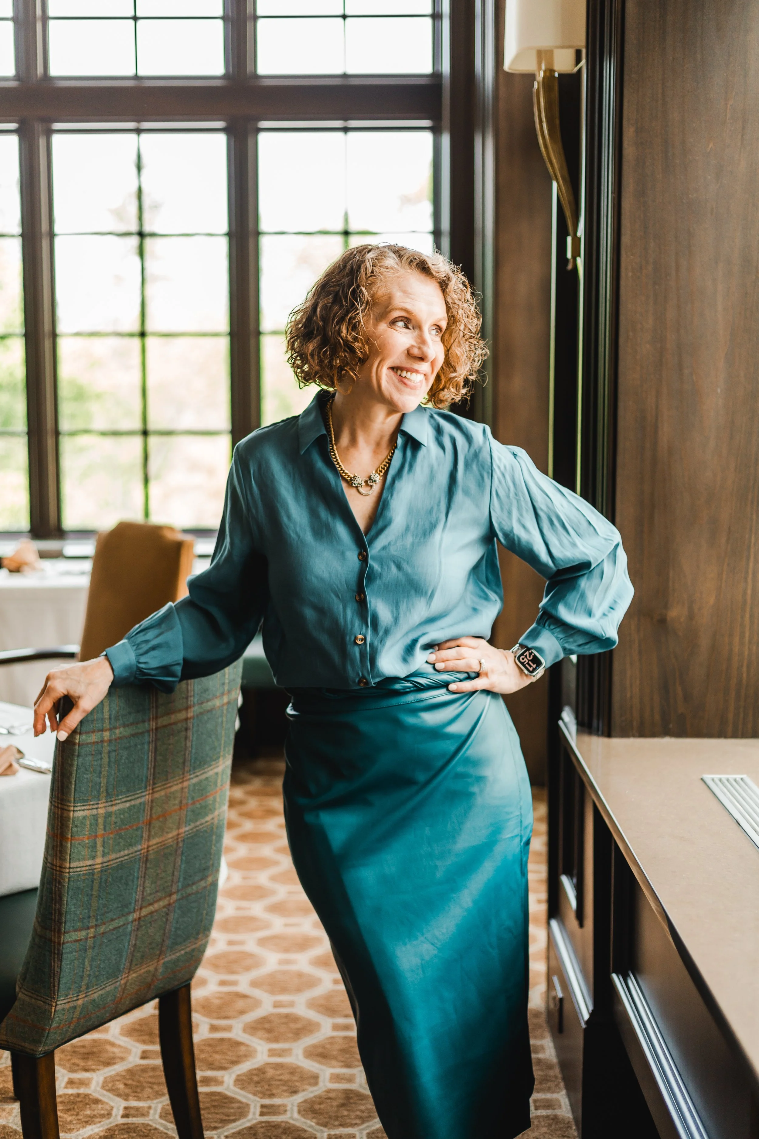 Woman smiling, wearing a teal blouse and skirt, standing in a restaurant with large windows and wooden accents.