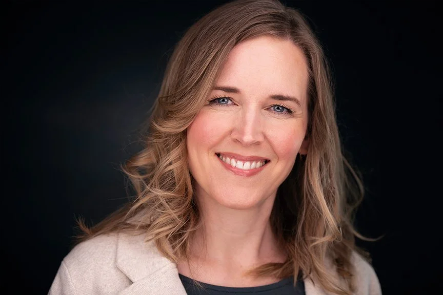 A woman with shoulder-length wavy blonde hair smiling against a dark background.