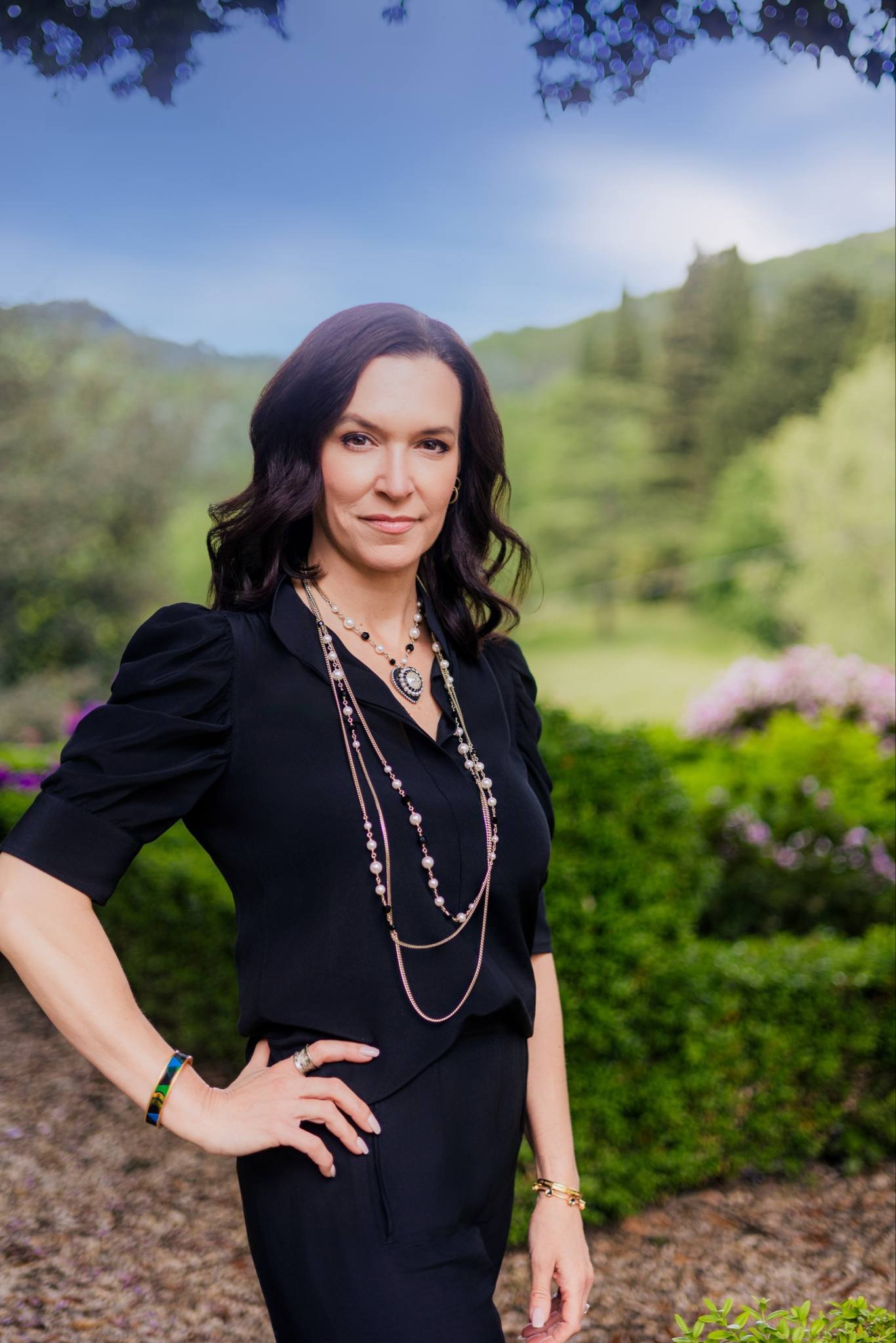 A woman with dark wavy hair wearing a black blouse and multiple layered necklaces standing outdoors in a lush green park with trees and pink flowers in the background.