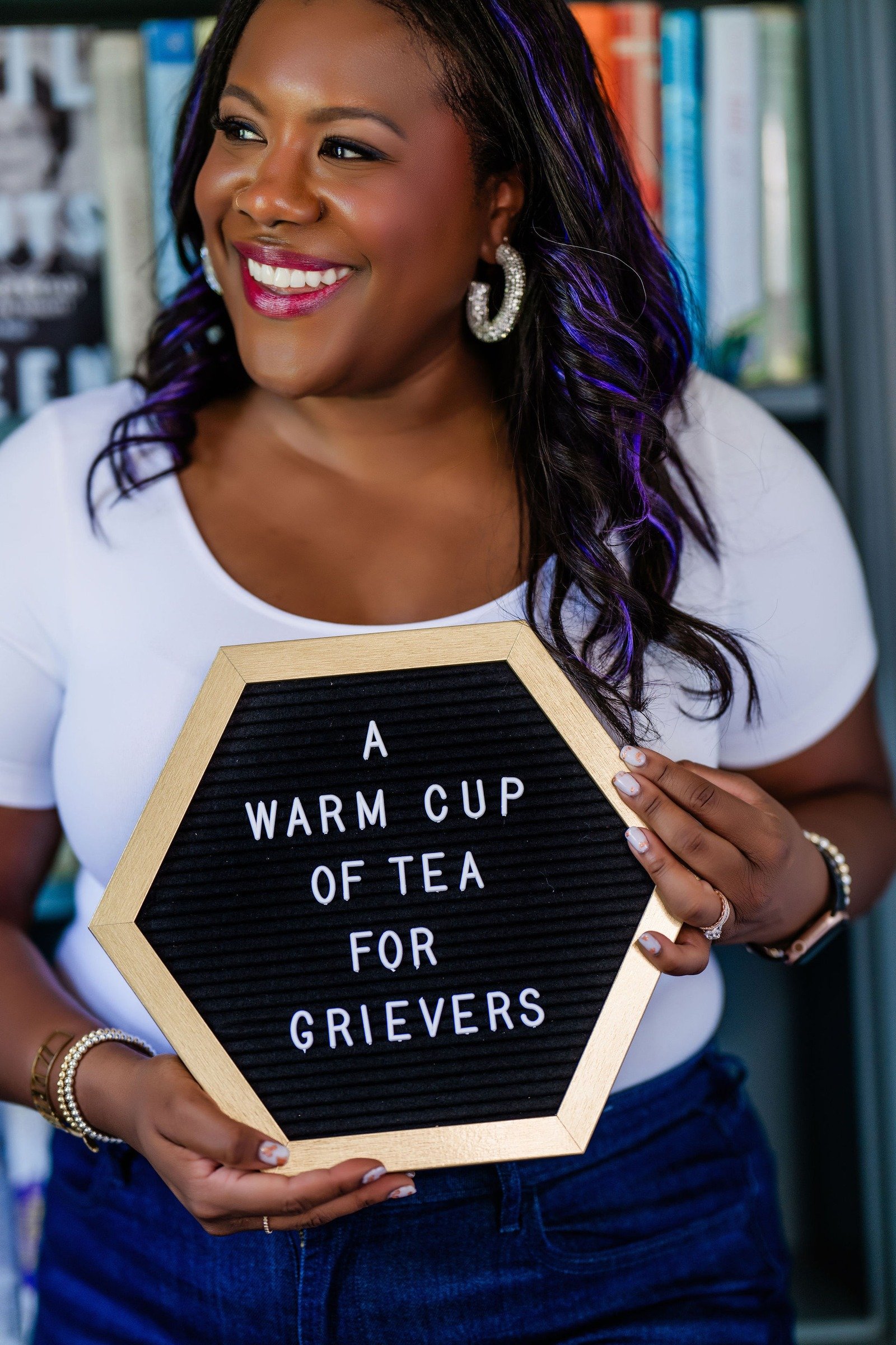 A woman with long dark hair, hoop earrings, and wearing a white top, smiling while holding a hexagonal black felt letter board with a wooden frame that reads, 'A WARM CUP OF TEA FOR GRIEVERS,' standing in front of a blurred bookshelf background.
