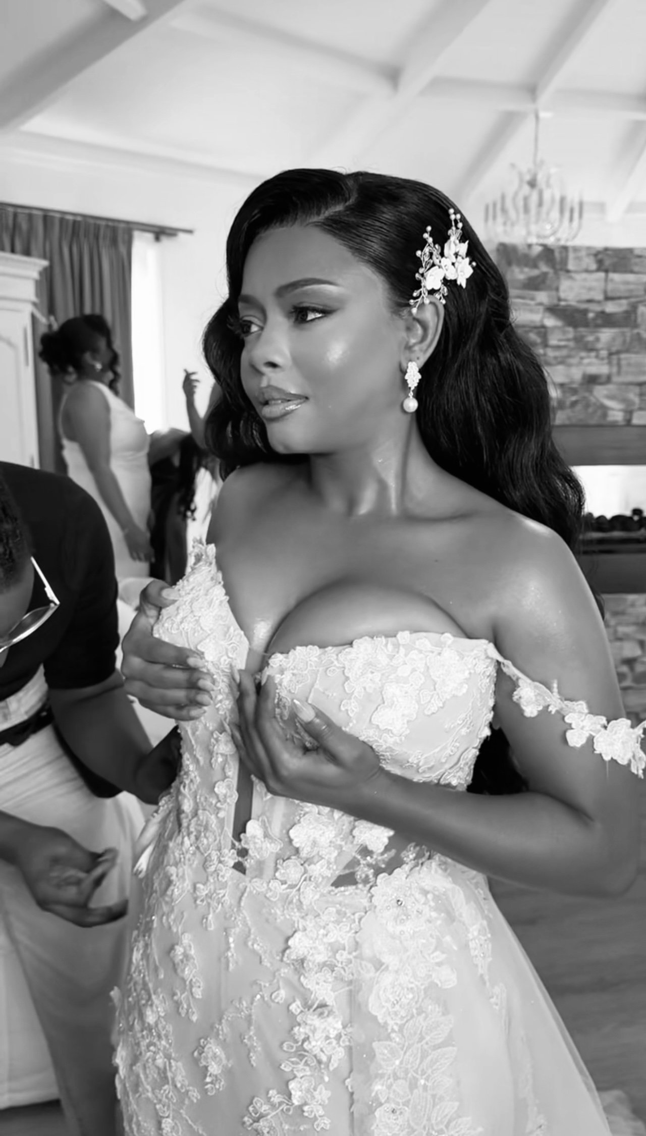 A bride in a wedding dress is adjusting her gown on her chest, with a woman in the background helping someone else. The bride is wearing ornate earrings and a floral hair accessory, and is posing indoors.