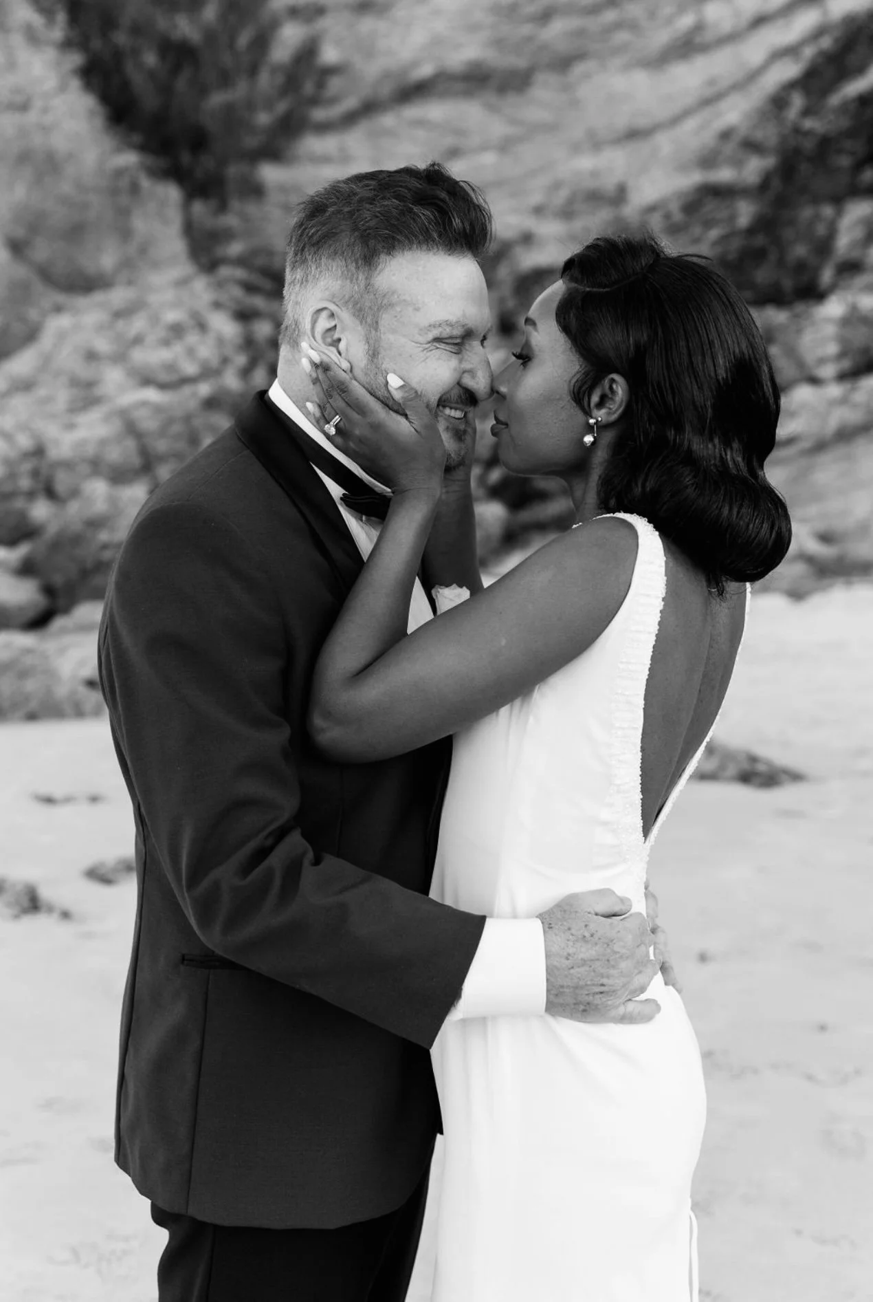 A black and white photo of a couple on their wedding day, kissing passionately with their eyes closed. The man is wearing a tuxedo and the woman is wearing a sleeveless wedding dress. They are on a beach with rock formations in the background.