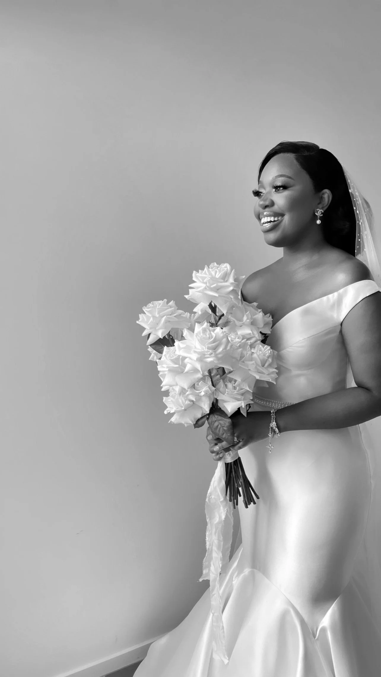 A smiling bride in a satin off-shoulder wedding dress, holding a bouquet of white roses, wearing earrings and a bracelet with a veil in her hair.