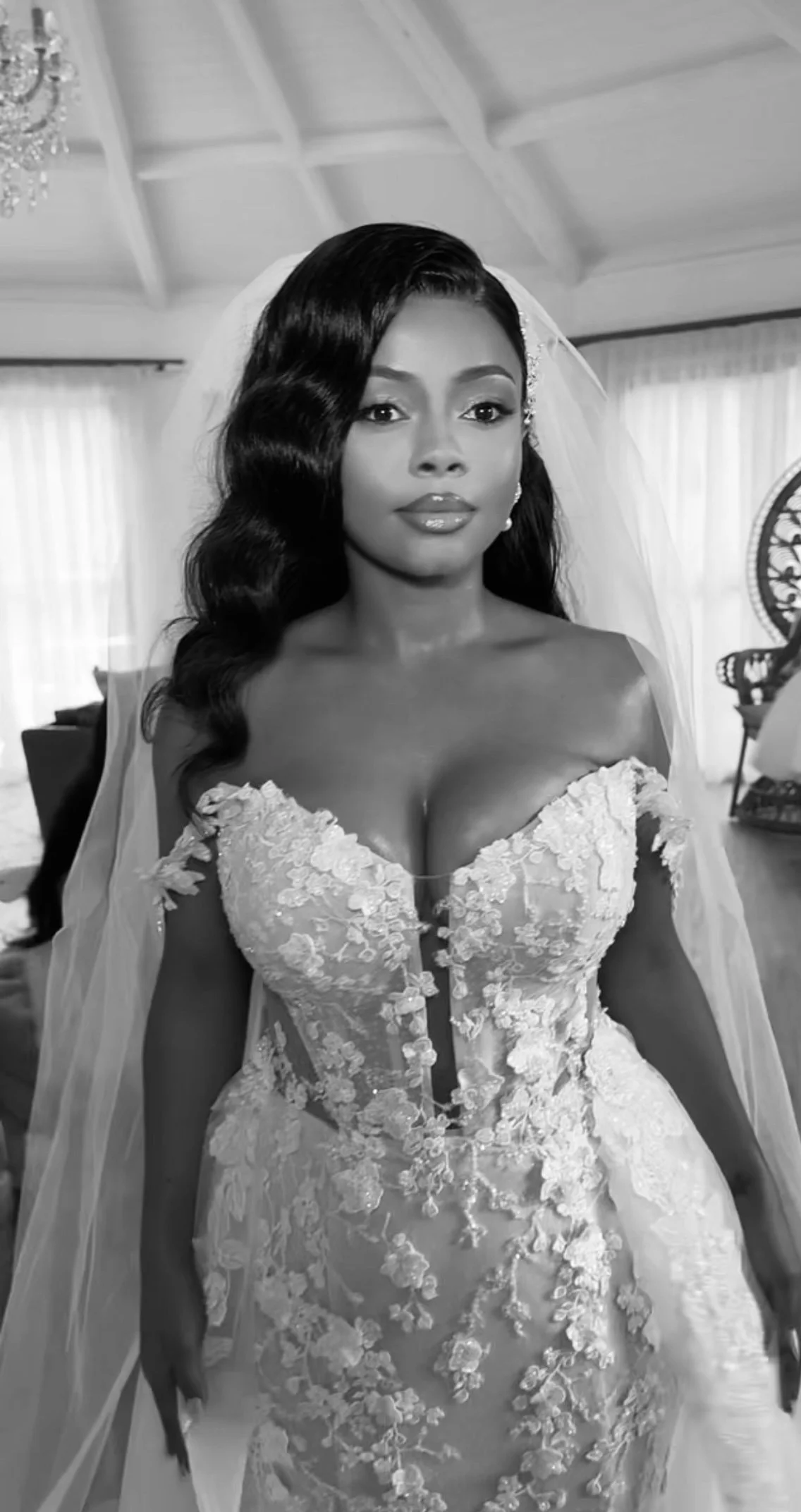 A bride in a lace wedding gown with off-the-shoulder sleeves, long veil, and styled hair in loose waves, standing in a well-lit room.