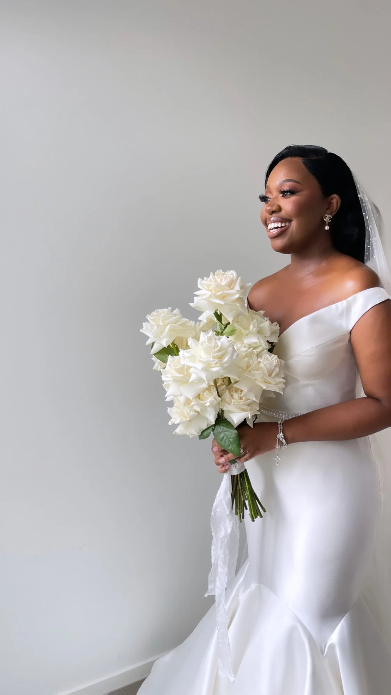 A bride in a white wedding dress holding a bouquet of white roses, standing against a plain gray wall.