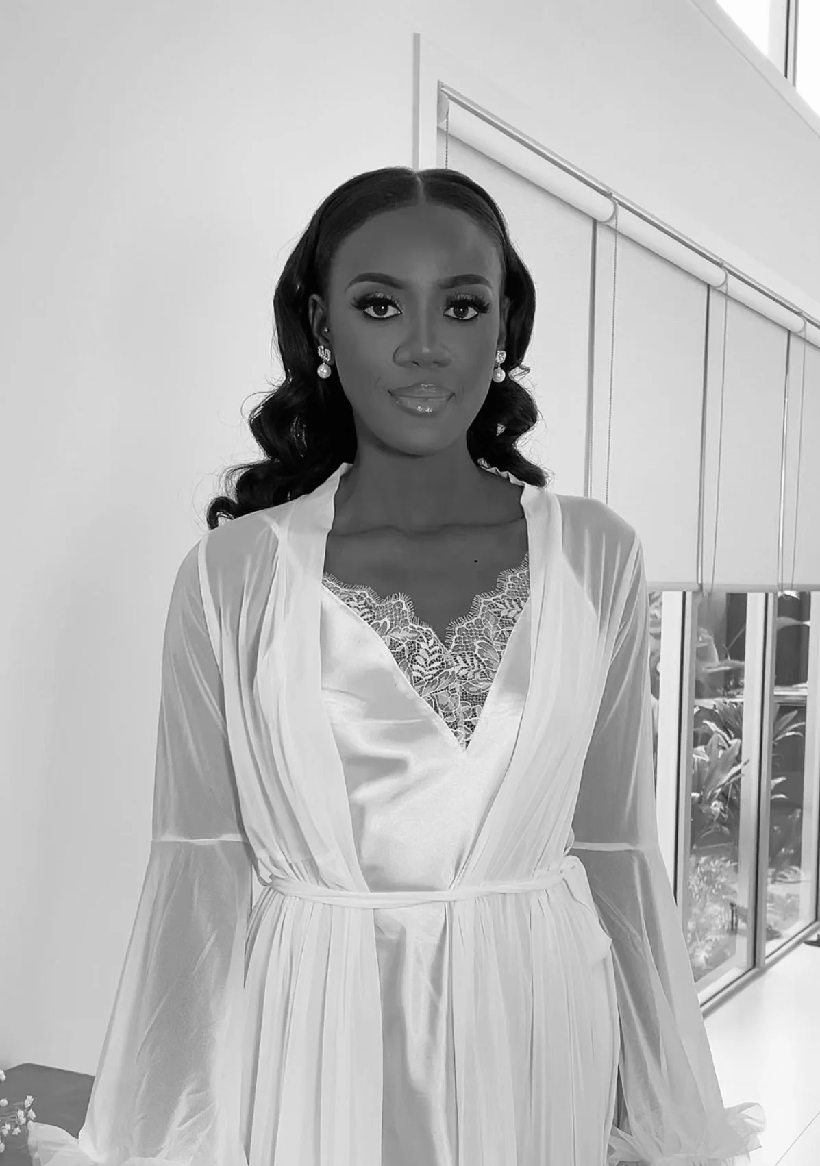 A woman wearing a light-colored robe with lace detail, standing indoors in front of a window and white wall.