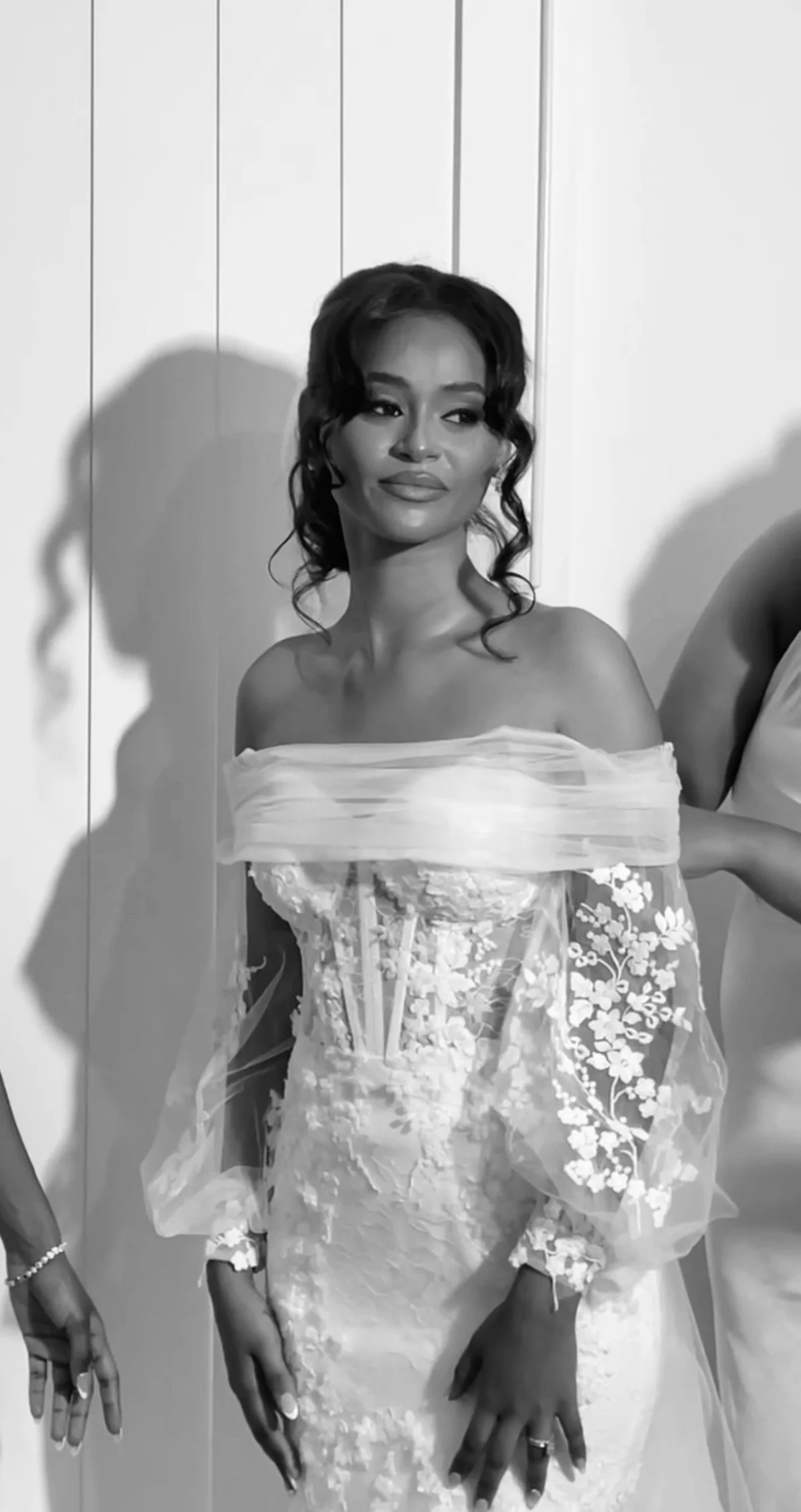 A woman in an elegant off-the-shoulder wedding dress with floral lace details, posing against a plain wall.