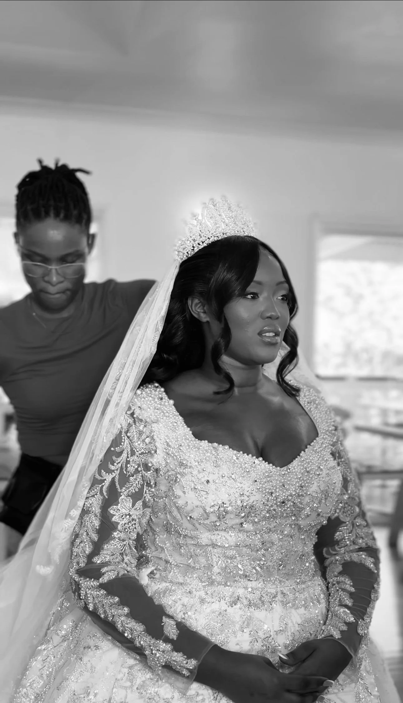 A woman dressed in a wedding gown with intricate beadwork, wearing a tiara and veil. Another woman stands behind her adjusting the gown in a bright room with windows.