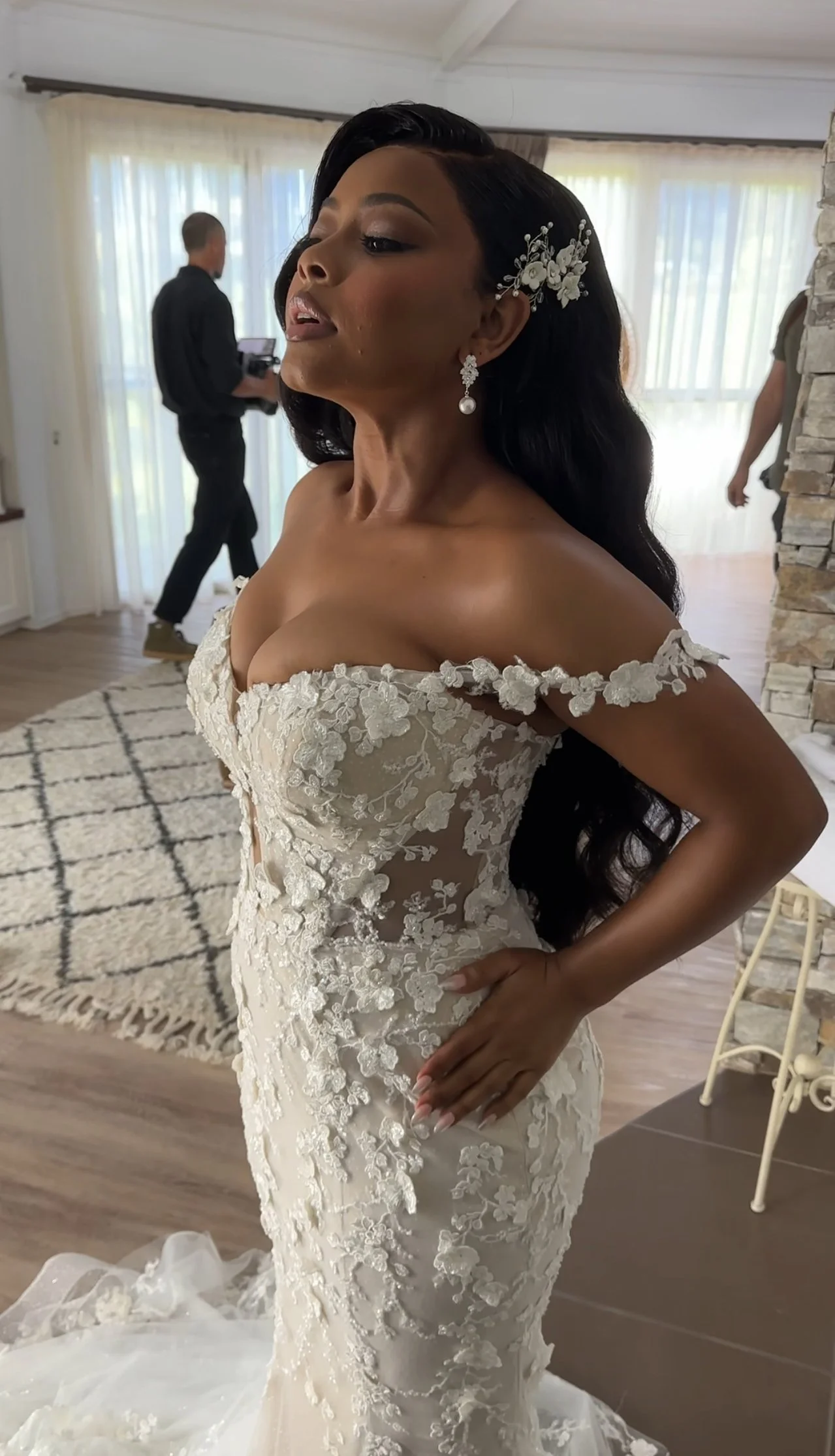 Bride in a wedding dress with off-the-shoulder floral lace details, wearing earrings and a floral hairpiece, inside a well-lit room with curtains and a patterned rug.