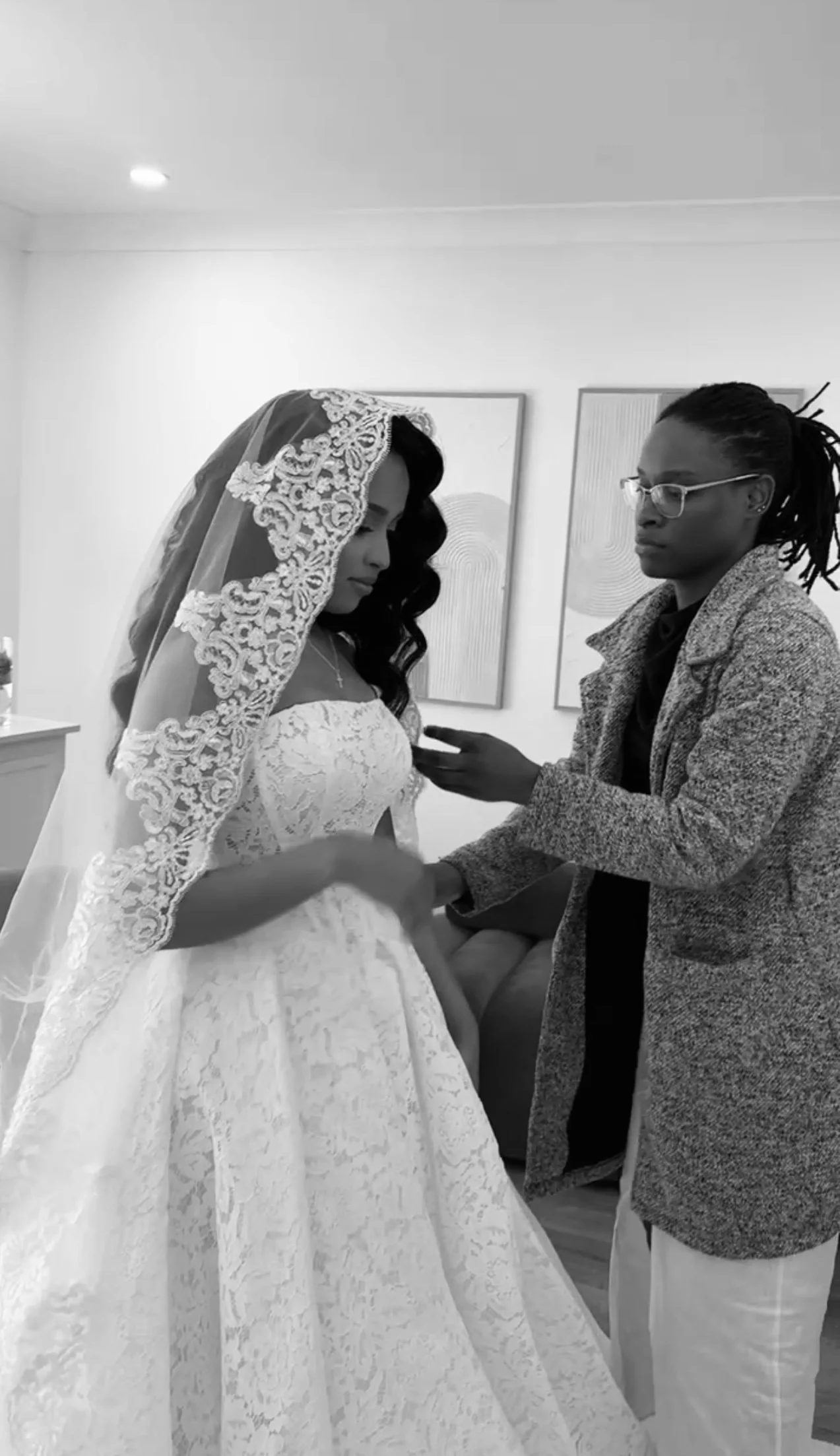 A bride in a lace wedding dress and veil standing in a room with artwork on the walls, while a woman adjusts her dress.