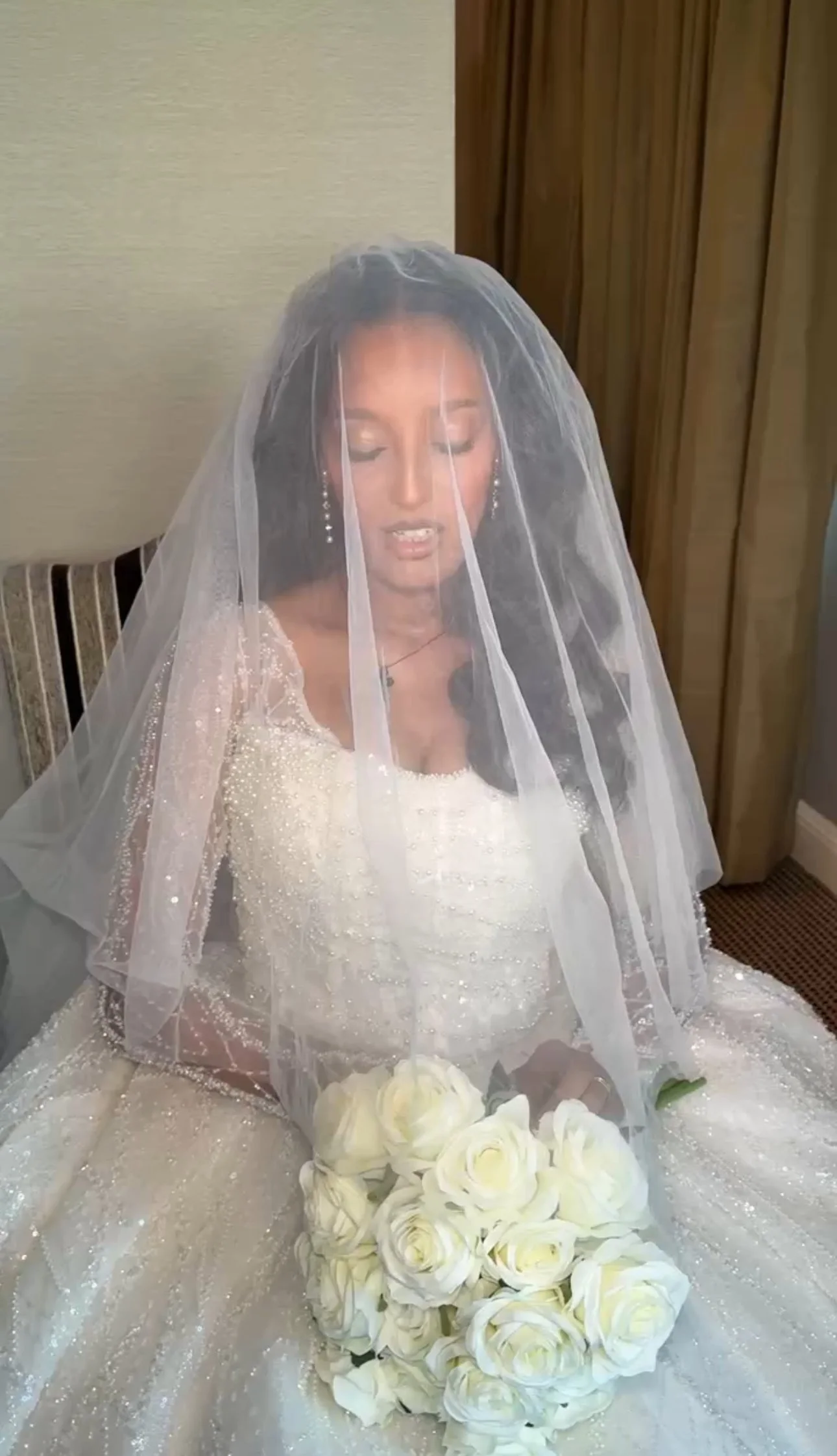 A bride with long dark hair in a wedding gown with a beaded bodice, wearing earrings and a necklace, holding a bouquet of white roses, sitting with a veil over her face in a room with wood-paneled walls and a beige wall.