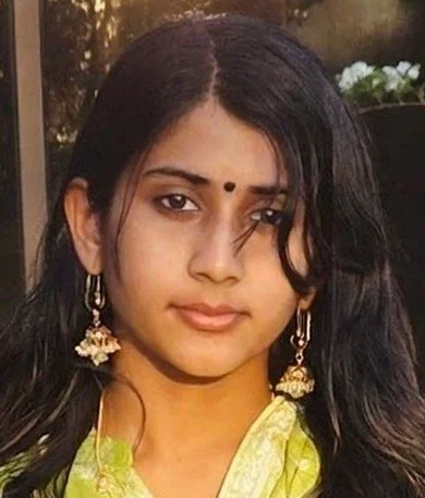 A woman with long dark hair and a black bindi on her forehead, wearing traditional earrings and a yellow outfit, outdoors in natural light.