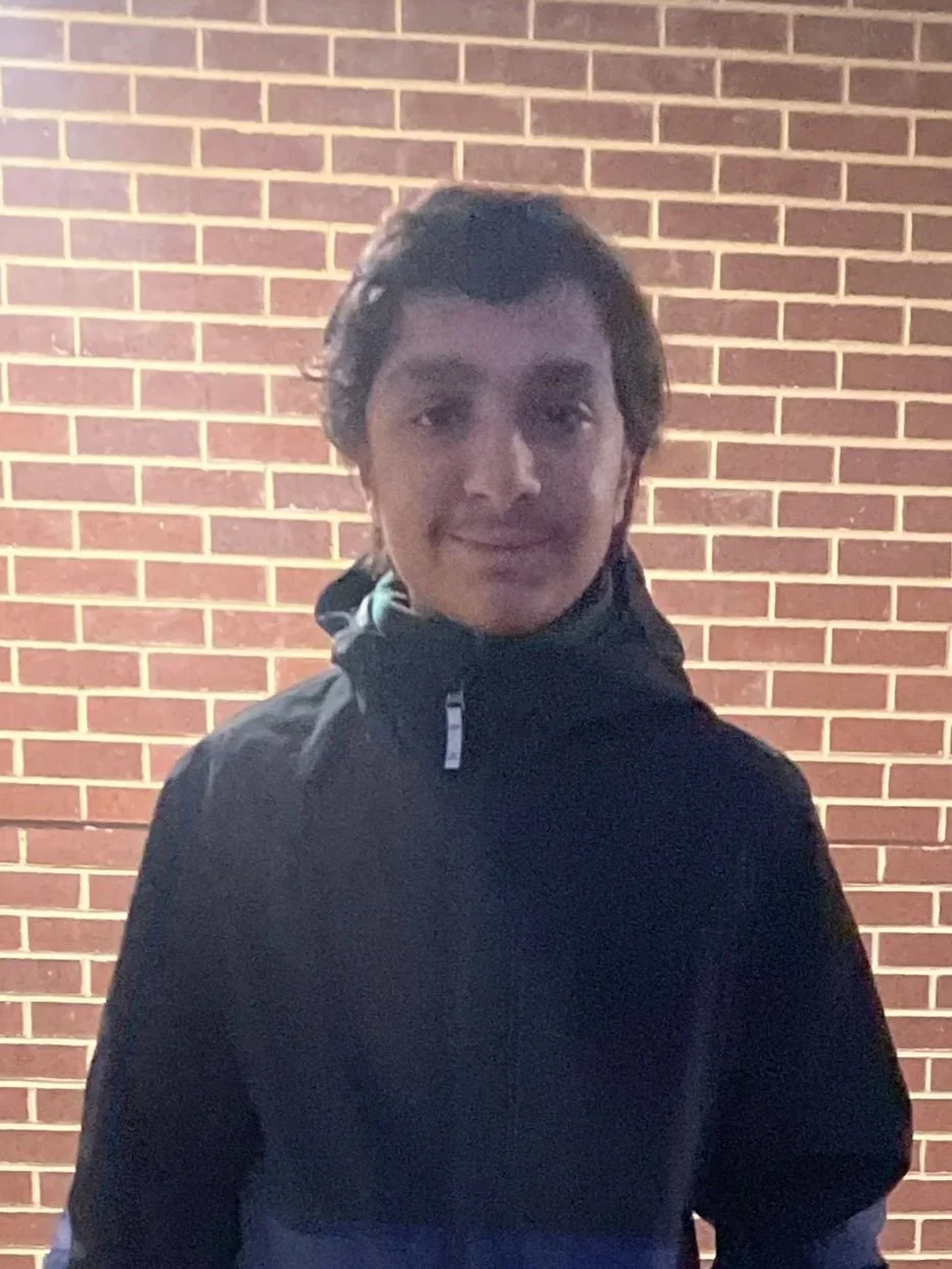 A young man with dark hair, wearing a dark jacket, standing in front of a red brick wall at night.