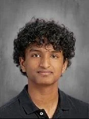 Portrait of a young man with curly black hair wearing a black collared shirt against a gray background.