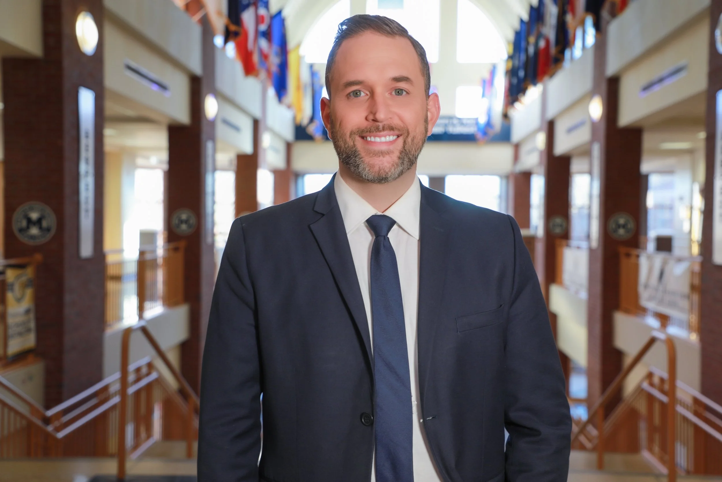 A smiling man in a navy suit with a tie standing inside a building with flags hanging from the ceiling.