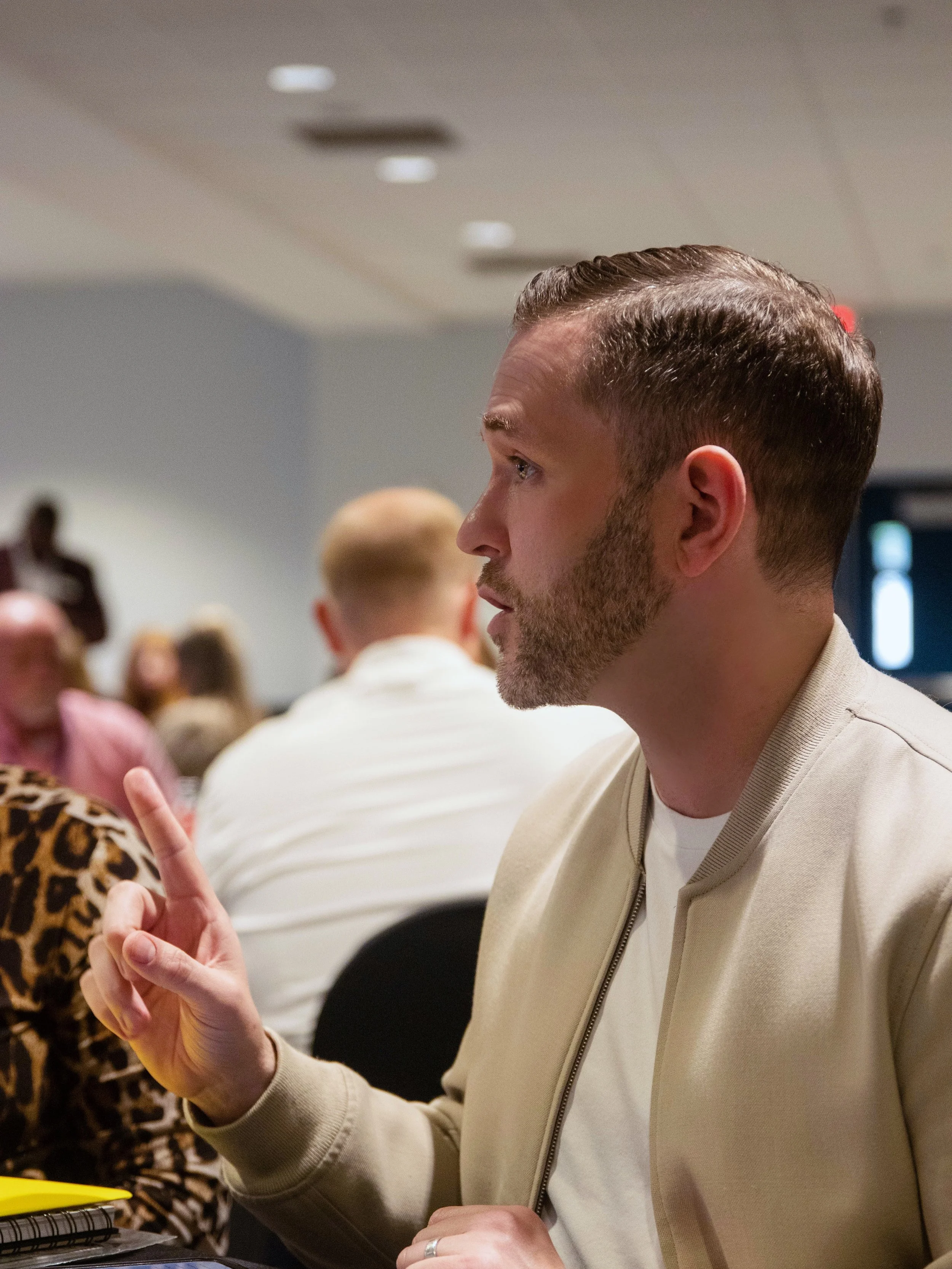 A man with short hair and a beard, wearing a beige jacket, is engaged in conversation at a group event in a conference room, with other people sitting and listening in the background.