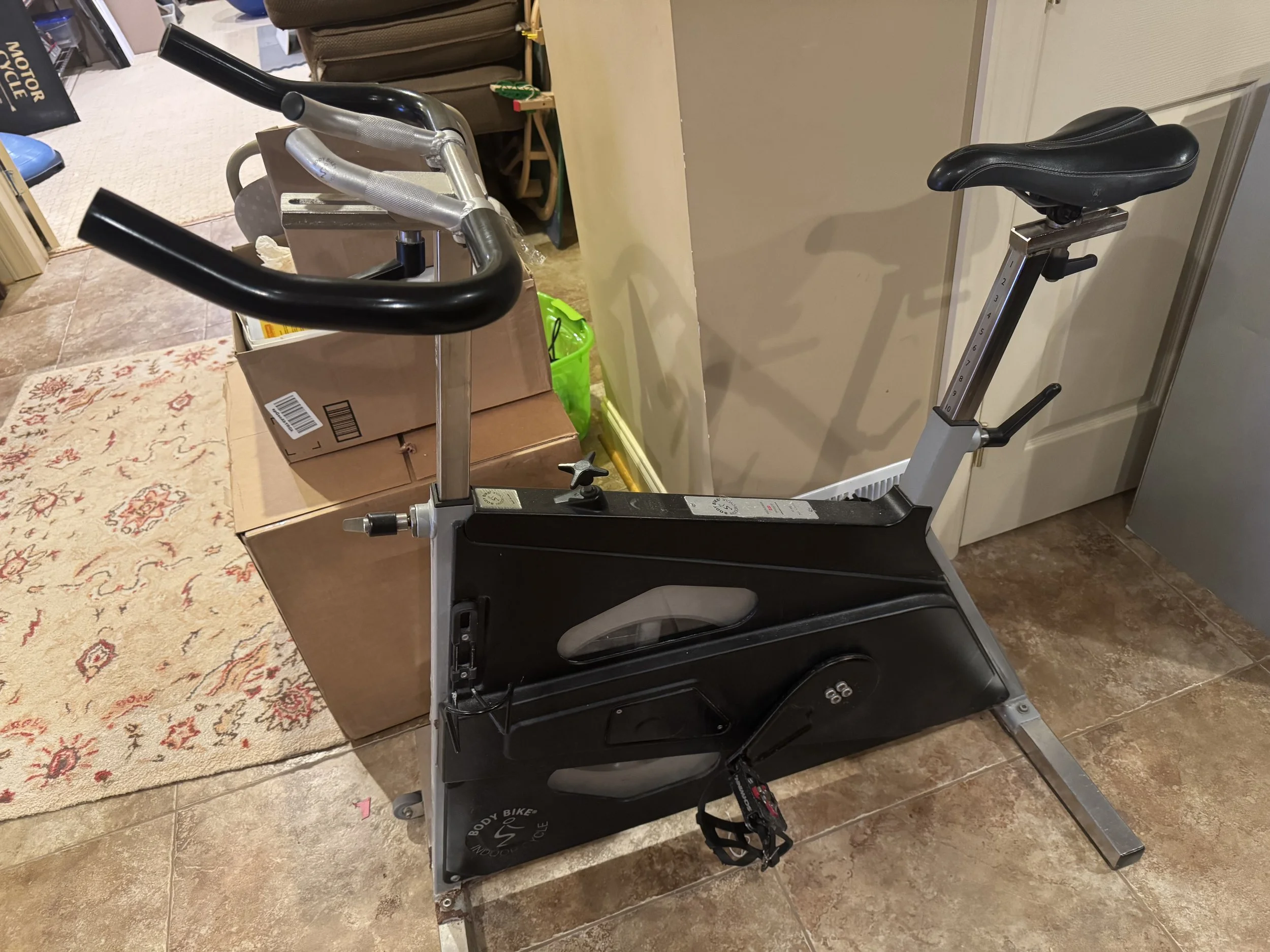 A stationary exercise bike indoors next to cardboard boxes and a green container.
