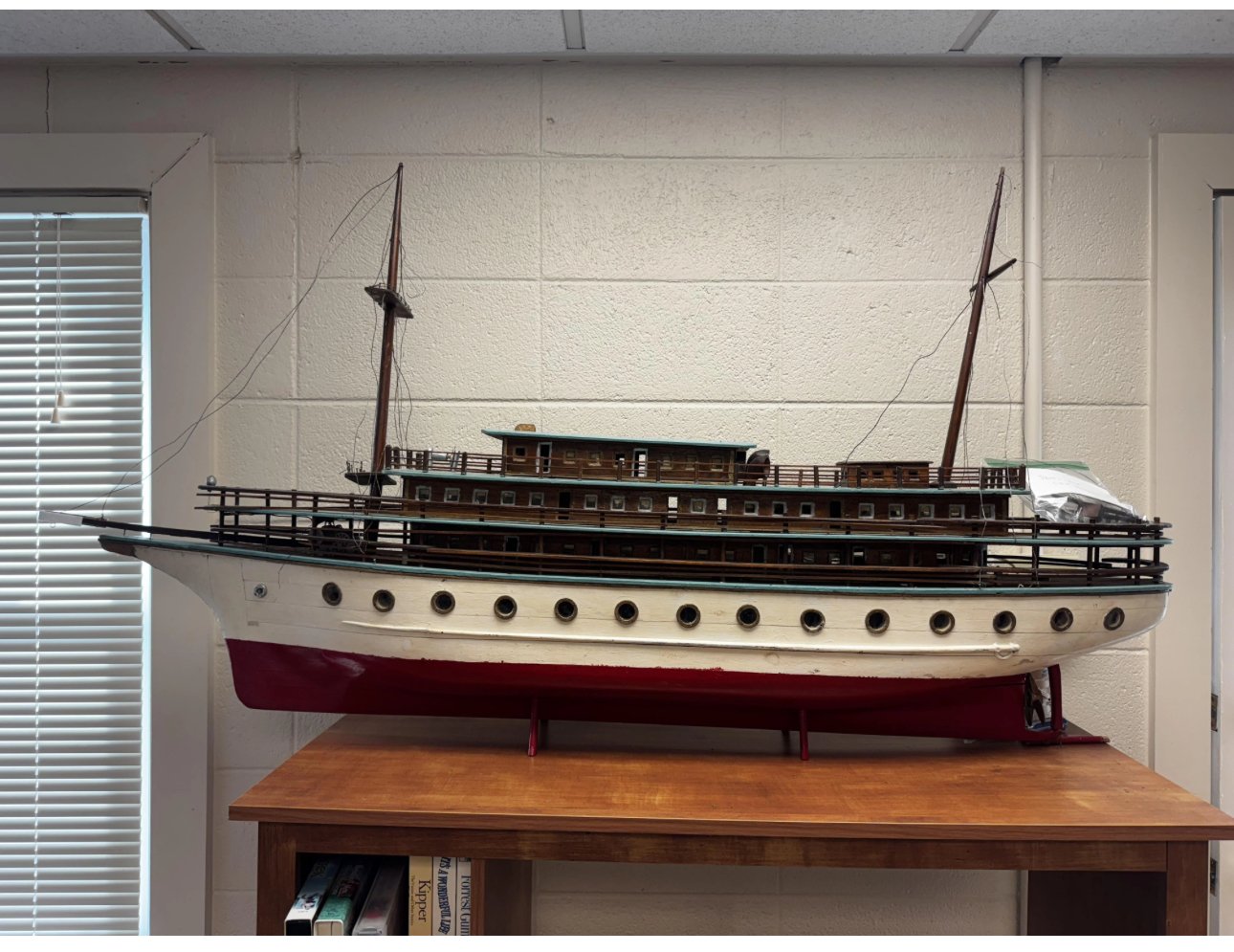 A detailed model of an old-style sailing ship displayed on a wooden table against a white brick wall, with a window with blinds on the left.