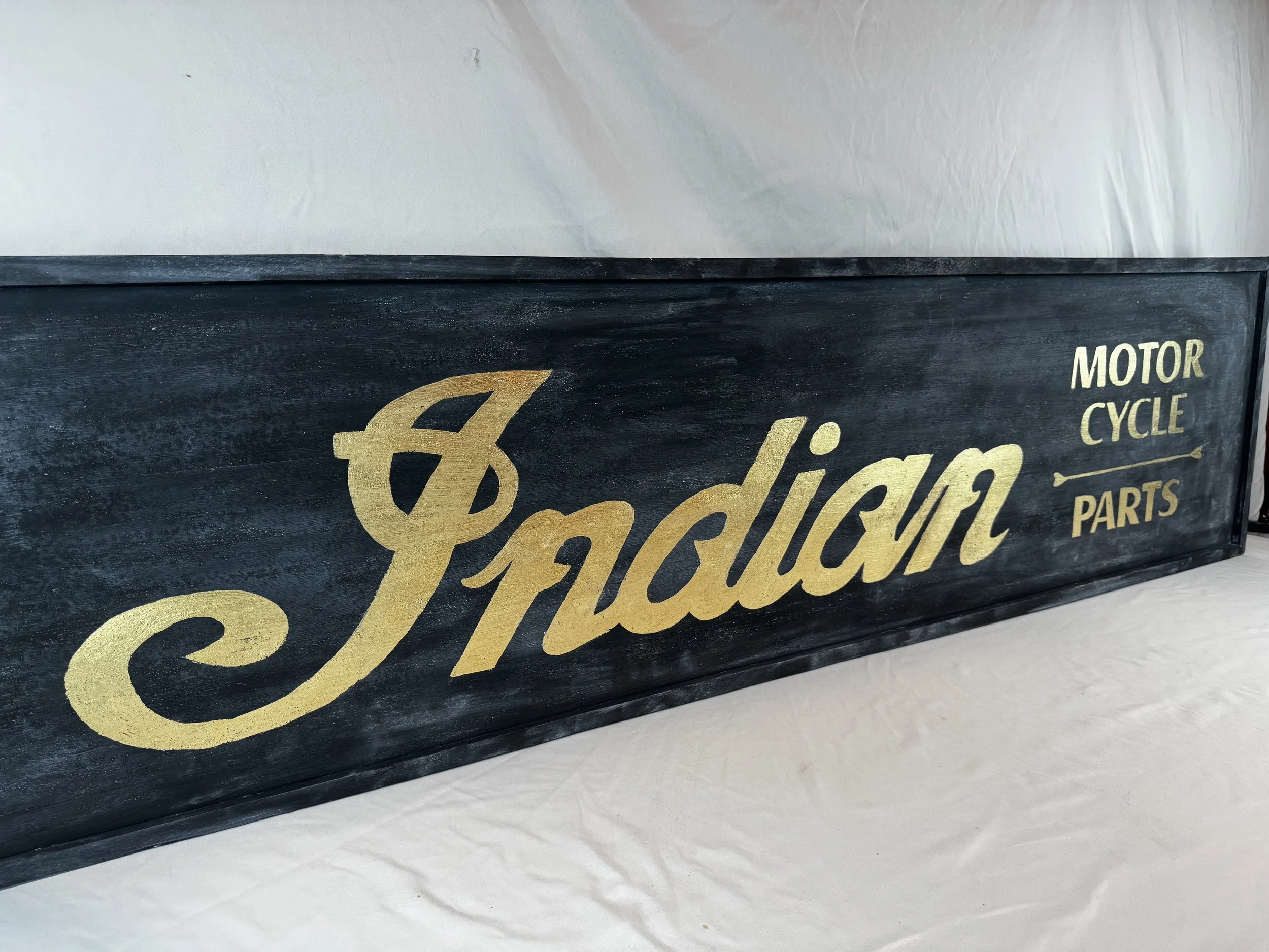 Black rectangular sign with smaltz and guild gold lettering reading "Indian Motor Cycle Parts".