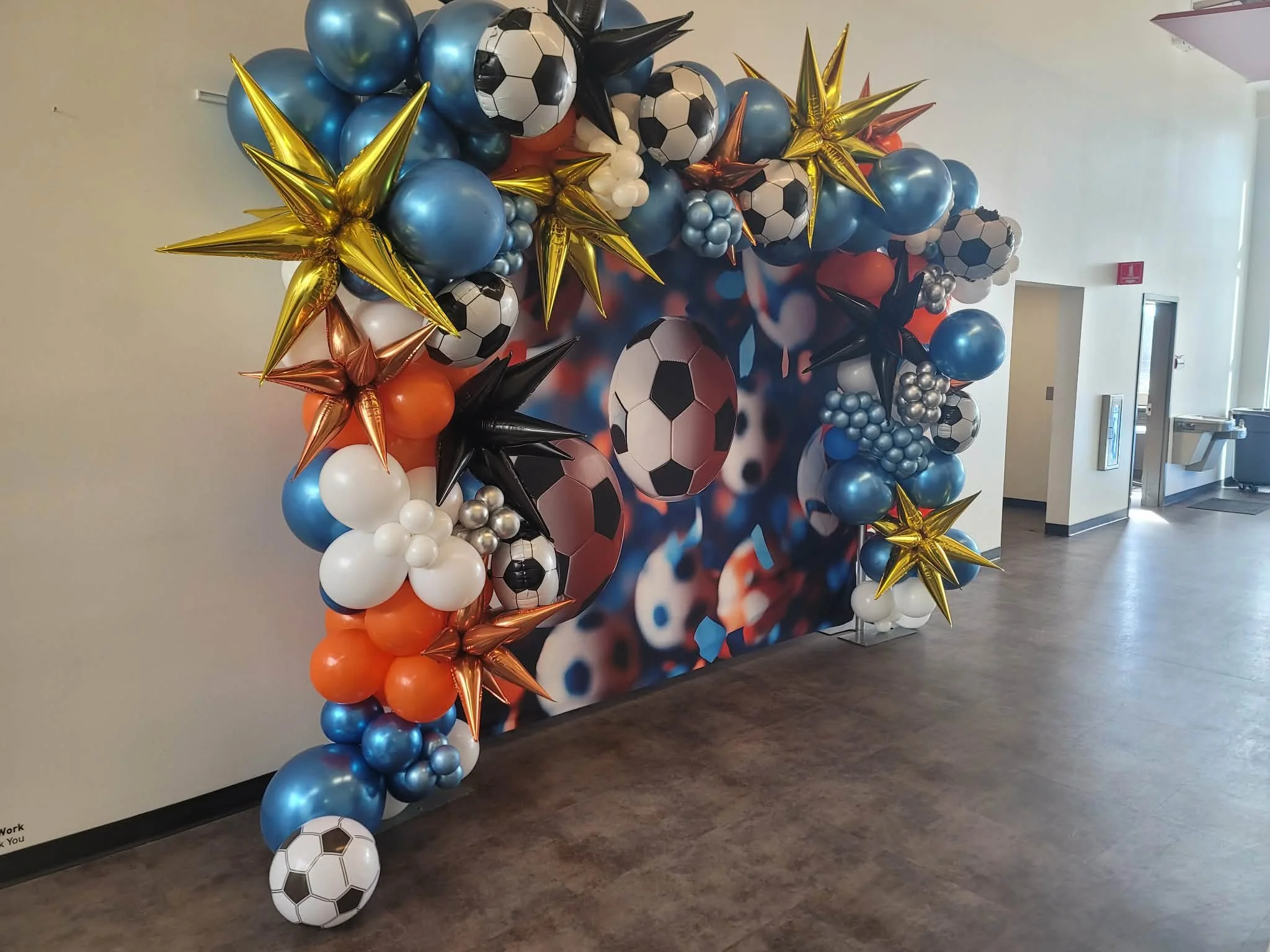 Colorful balloon arch featuring blue, orange, white, black, and metallic gold, silver, and rose gold balloons with soccer ball shapes, set against a backdrop with soccer-themed images.