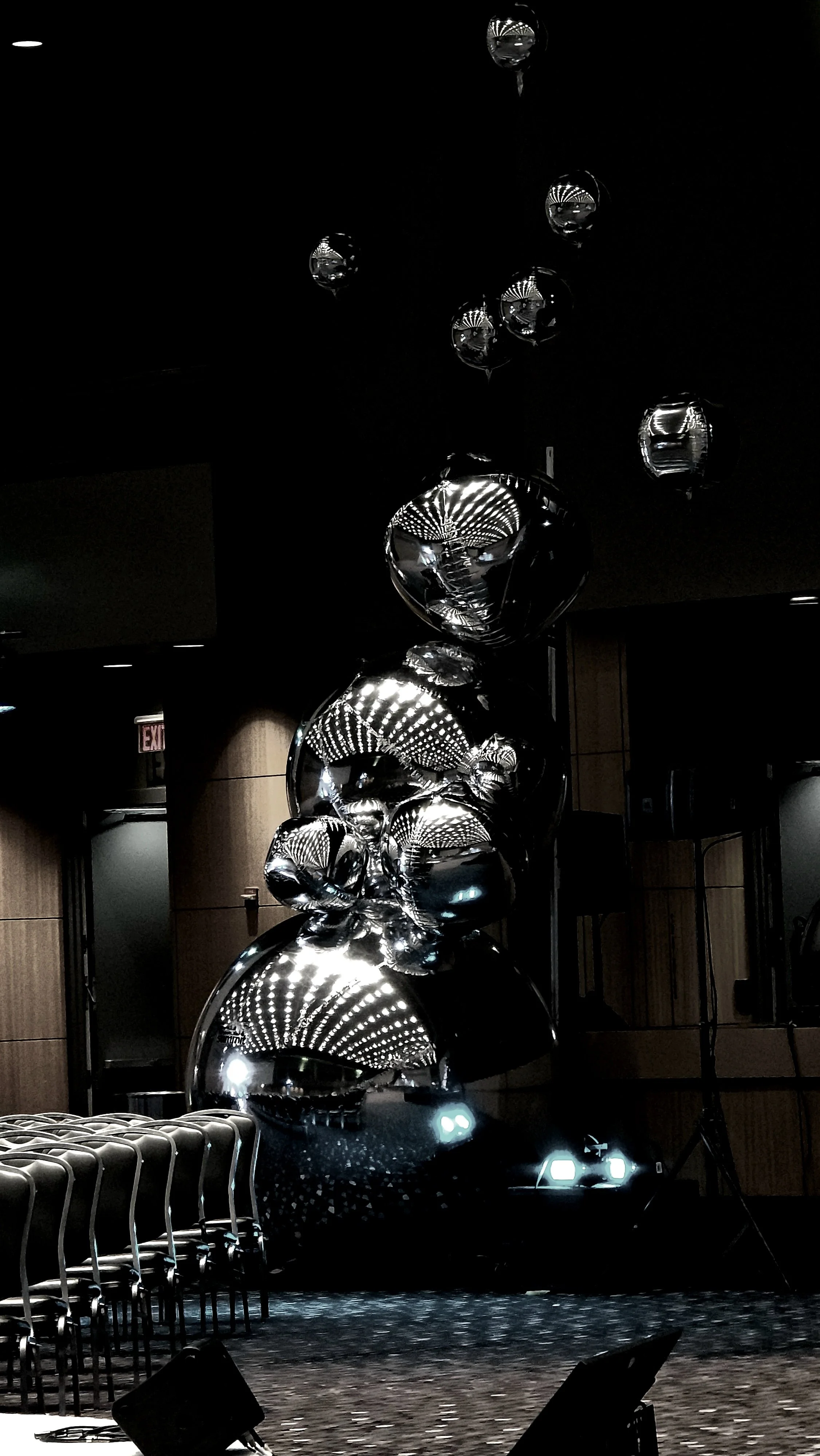 An art installation of reflective, metallic balloons stacked together in a dark room, with a mirror-like surface creating distorted reflections of the surroundings.