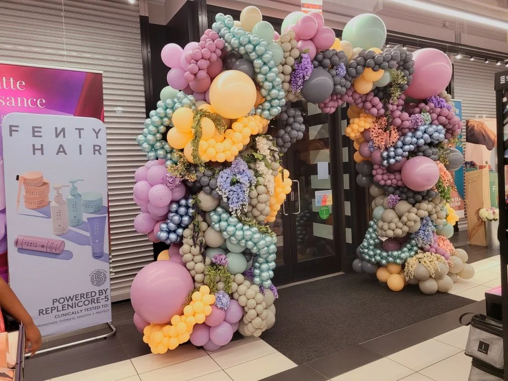Colorful balloon arch entrance with pastel and metallic balloons in purple, pink, yellow, blue, gray, and cream, decorated with flowers, at a shopping mall.