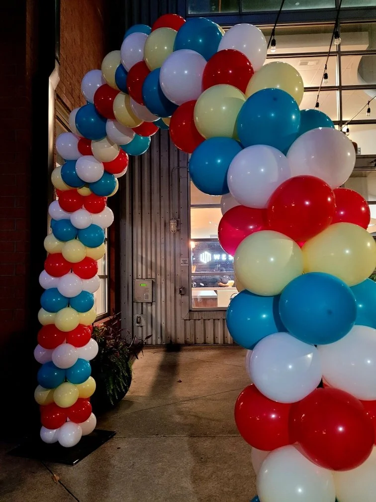 Colorful balloon arch made of red, white, blue, and yellow balloons at the entrance of a building.