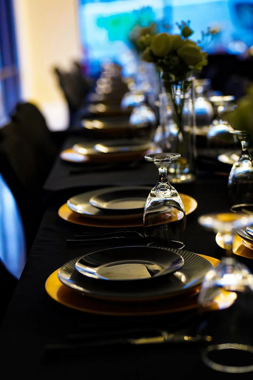 A formal dining table set with stacked plates, upside-down wine glasses, and floral centerpieces in vases, with a dark tablecloth and chairs along the sides.