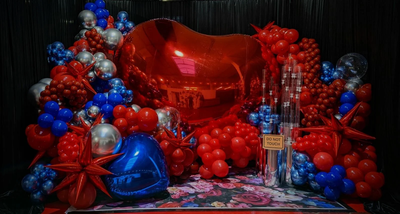 Decorative display with red, blue, and silver balloons surrounding a large red heart-shaped balloon in the center, with a sign that reads 'DO NOT TOUCH' and a floral-patterned floor.