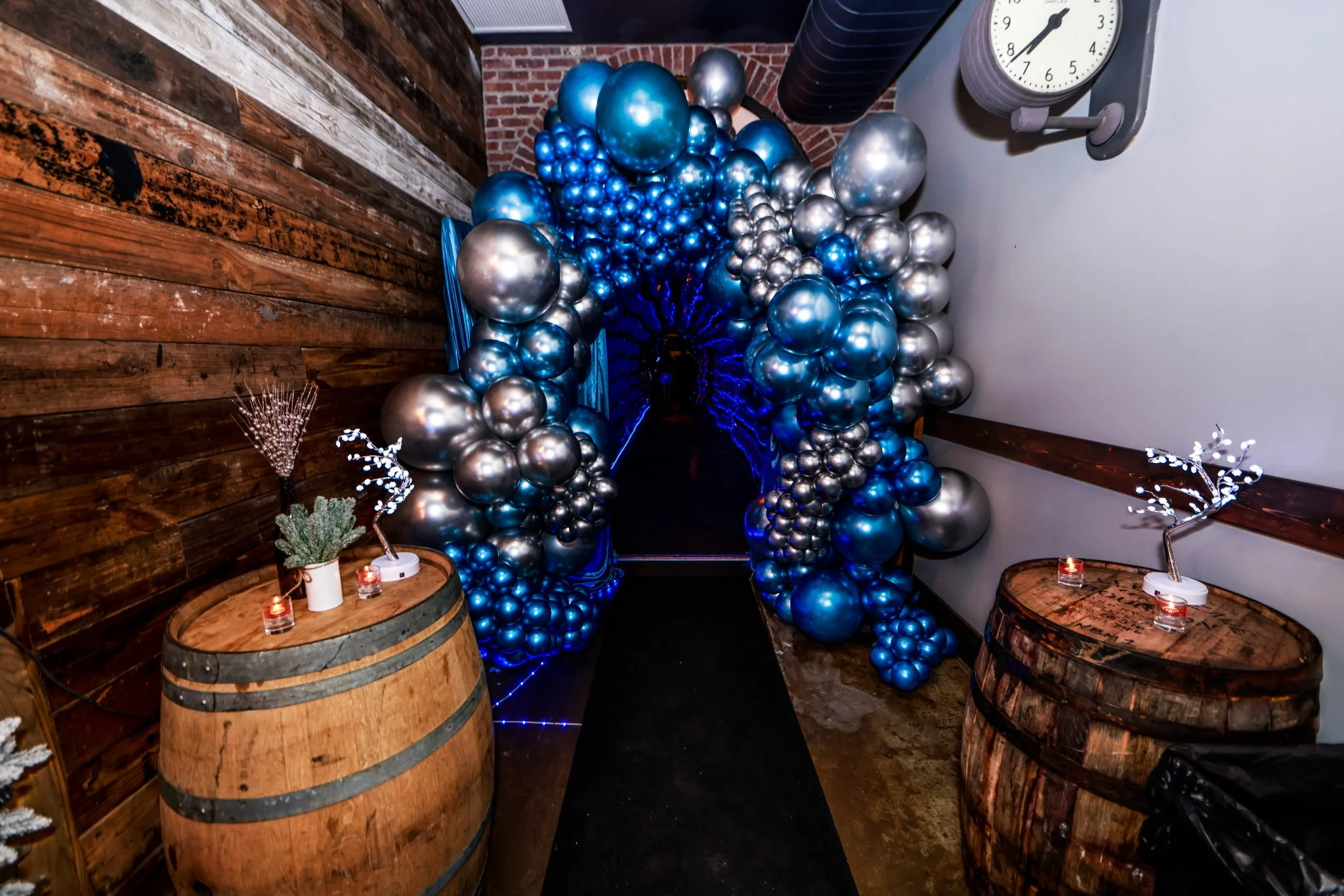 Decorative balloon arch with blue and silver balloons, flanked by wooden barrels with candles and small potted plants, in an indoor setting with rustic wood and brick walls.