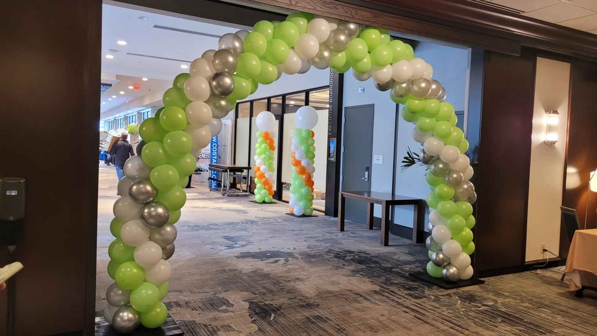Balloon arch made of green, white, silver, and gold balloons at entrance to an indoor event space with decorated columns in the background.