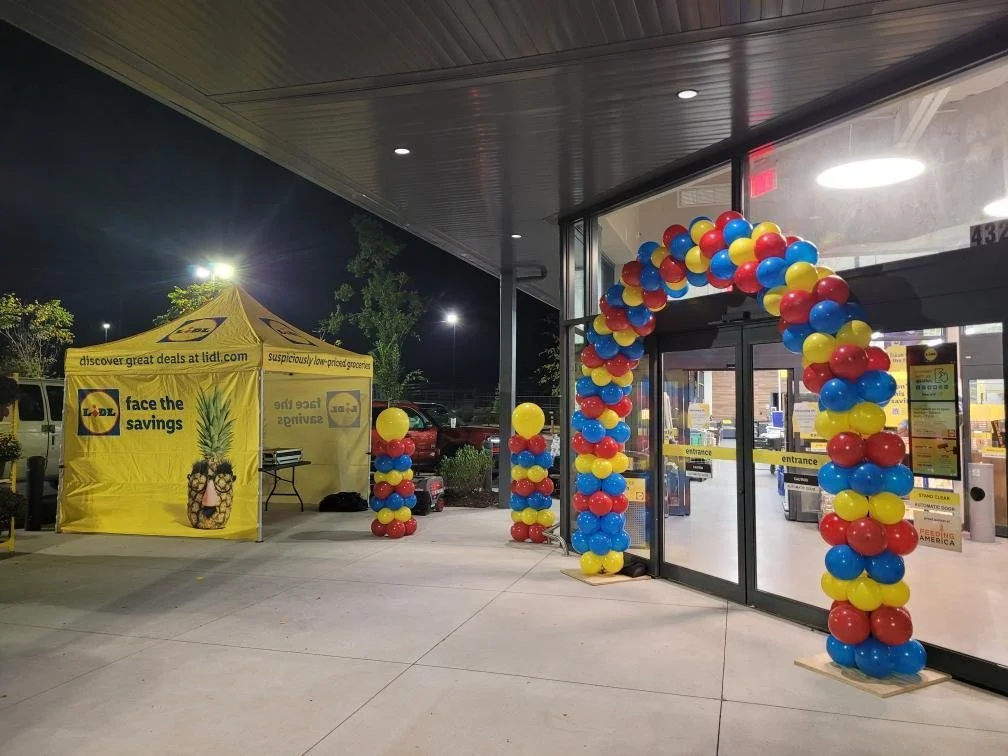 Decorated entrance of a store with a balloon arch made of red, blue, and yellow balloons and a yellow tent with the Lidl logo and pineapple graphic, indicating a promotional event.