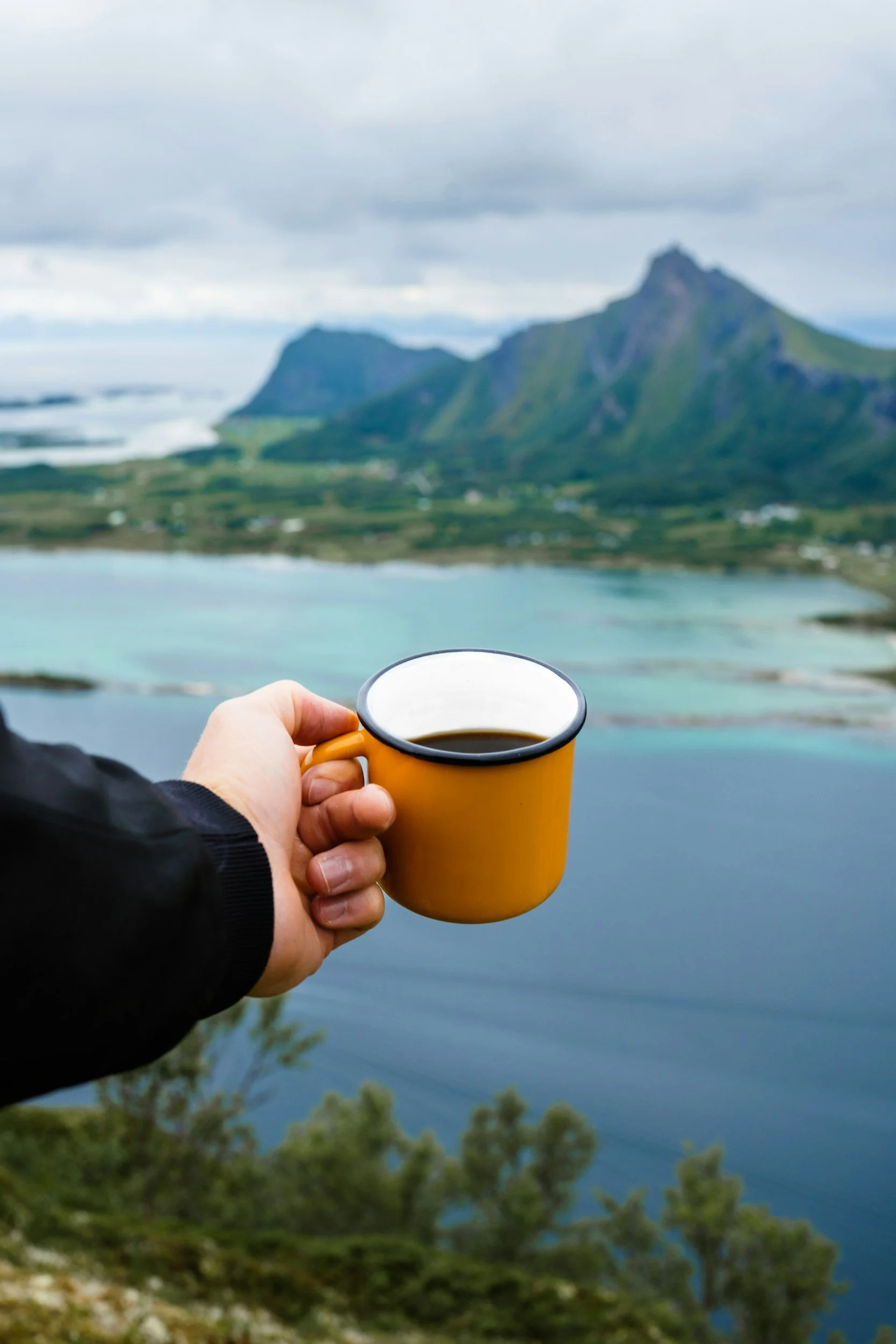 Person holding a yellow mug of coffee or tea with a mountainous landscape and a body of water in the background.