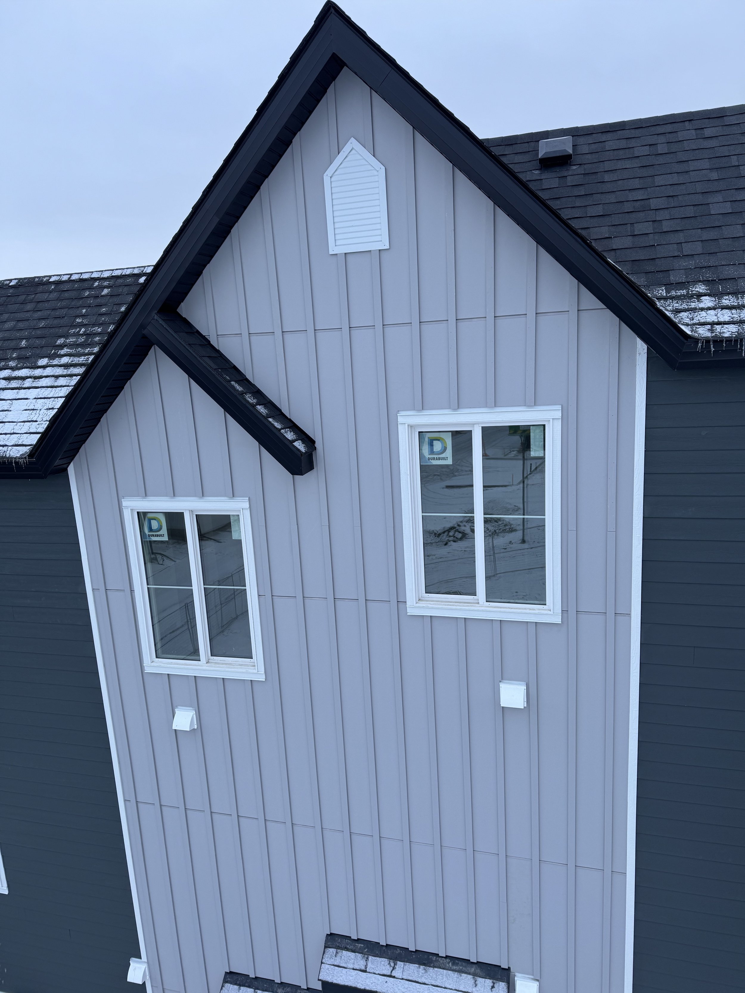 Close-up of a modern house's exterior corner, showing gray vertical siding, two double-hung windows, black roof, and small white vent on the gable.