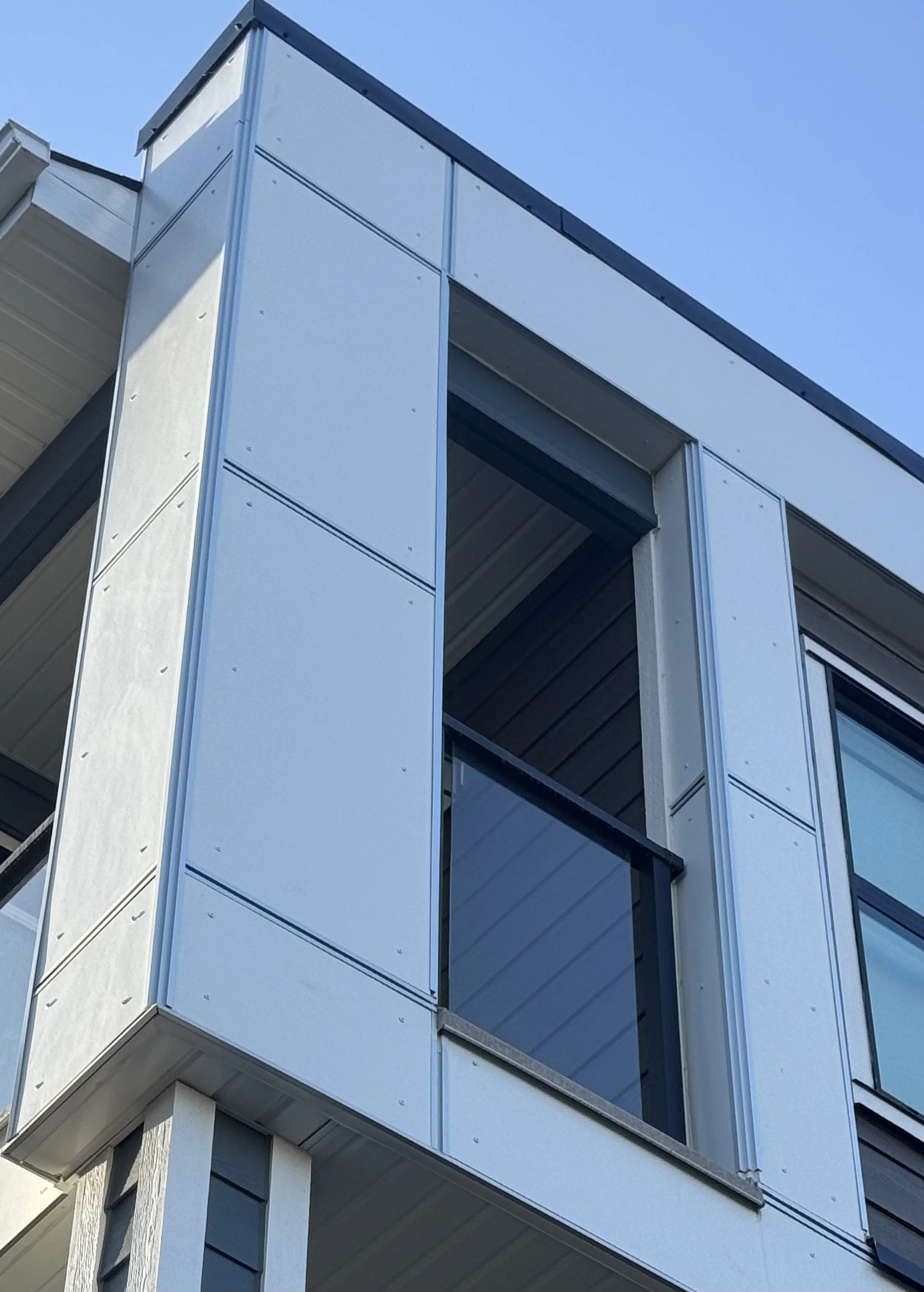 Close-up of a modern building's exterior corner with metal siding and a large window, under a clear blue sky.