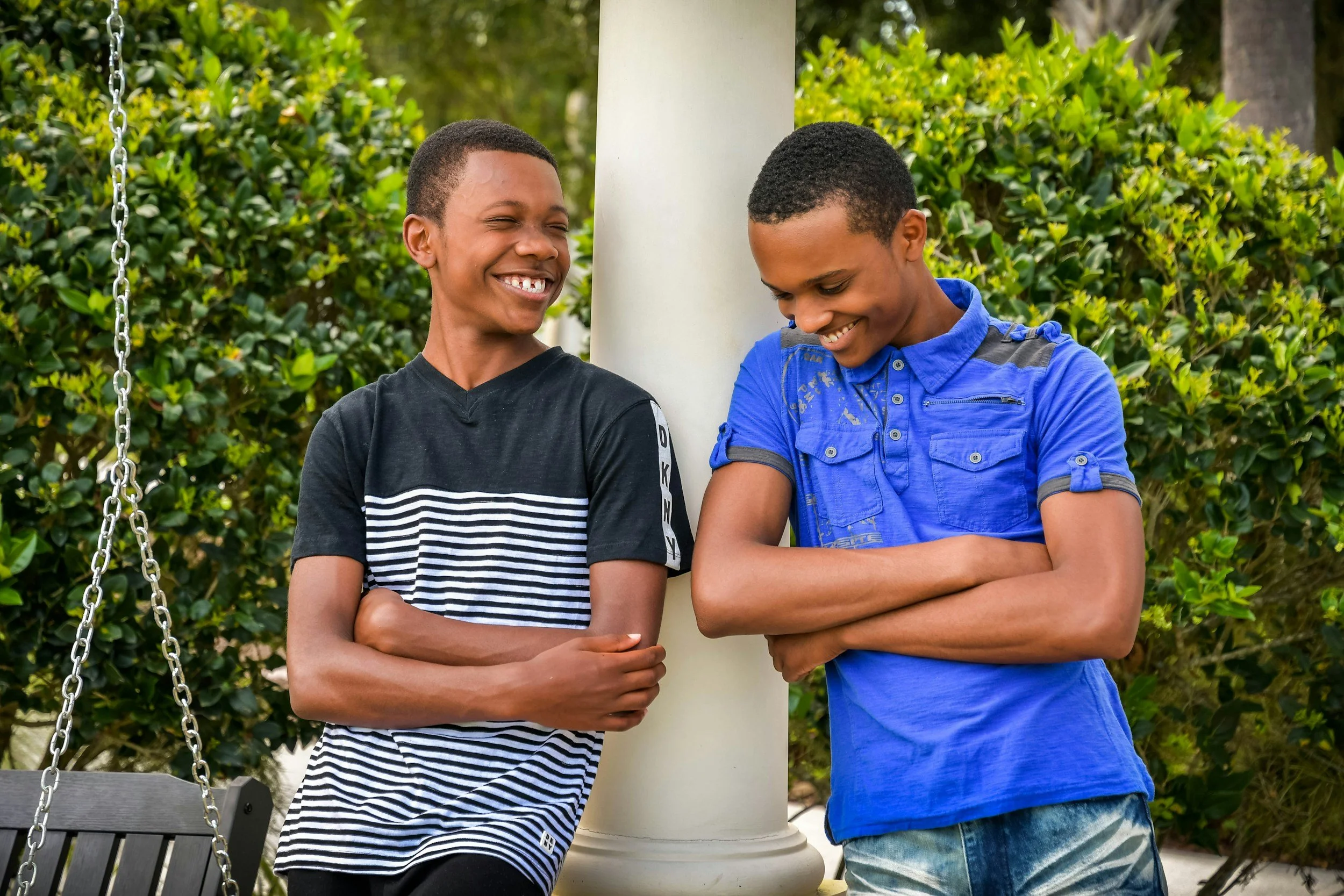 Two boys standing outside near a white post, smiling and looking at each other, with green bushes and a metal swing nearby.